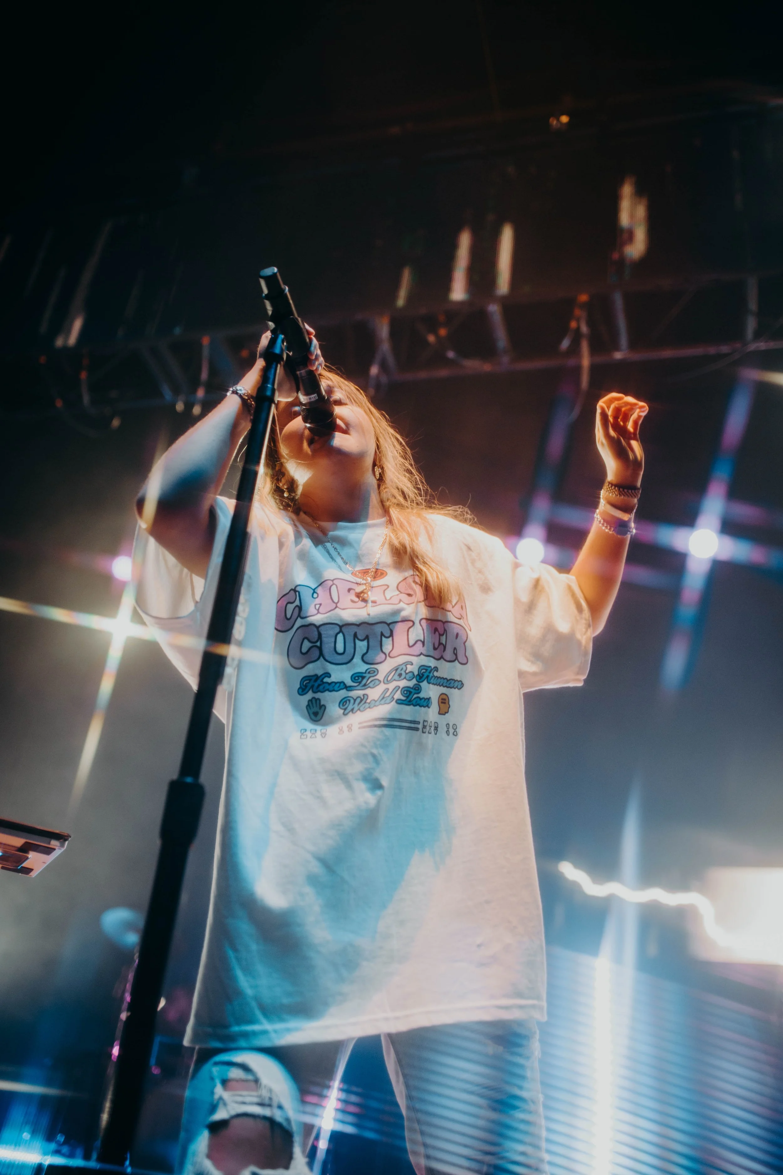 Chelsea Cutler performing on stage, holding a microphone, wearing a white oversized T-shirt with colorful text and graphics, and light-colored ripped jeans, with stage lights in the background in Detroit, Michigan.