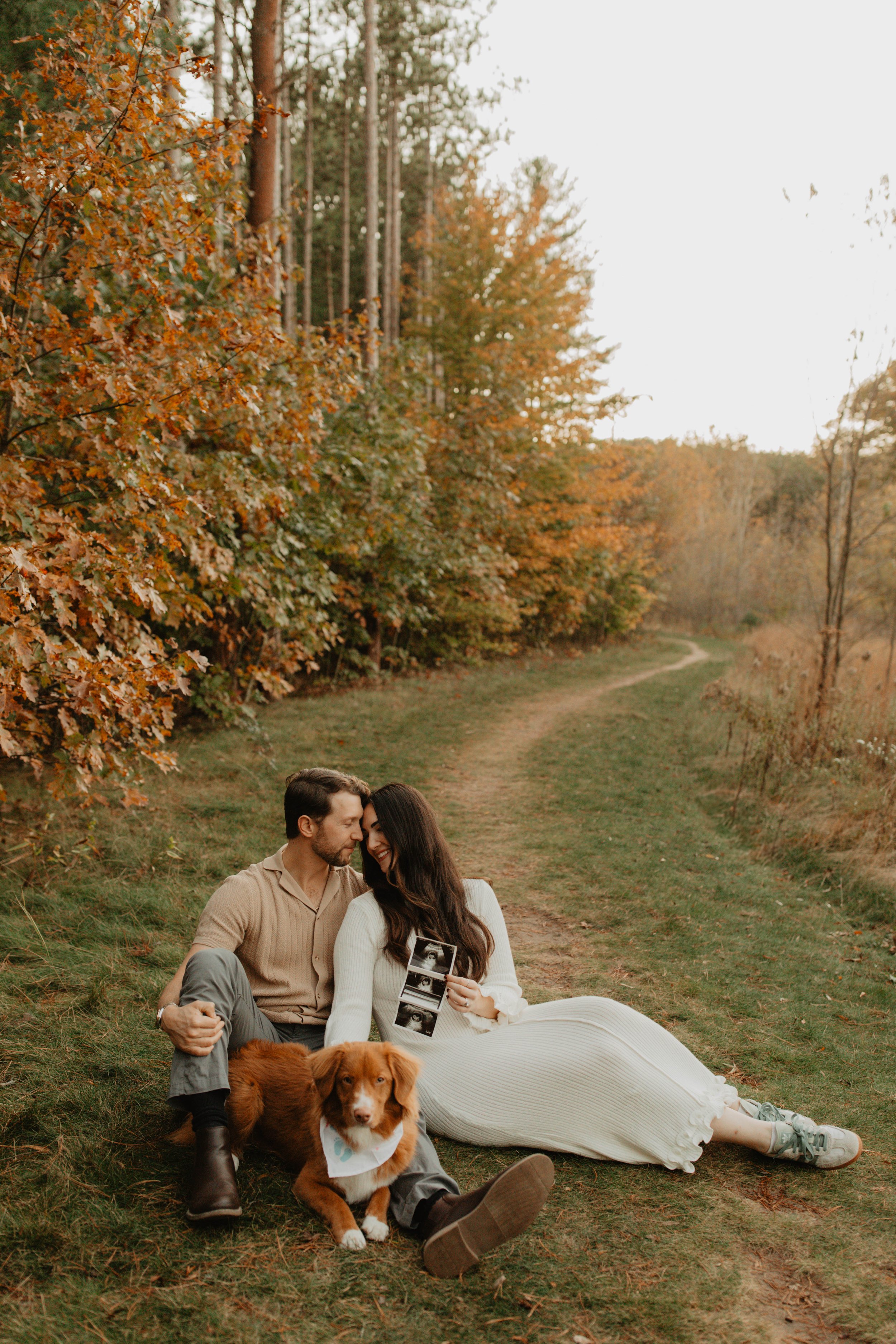 A couple sitting on a grassy path in autumn, with their dog and holding ultrasound pictures.