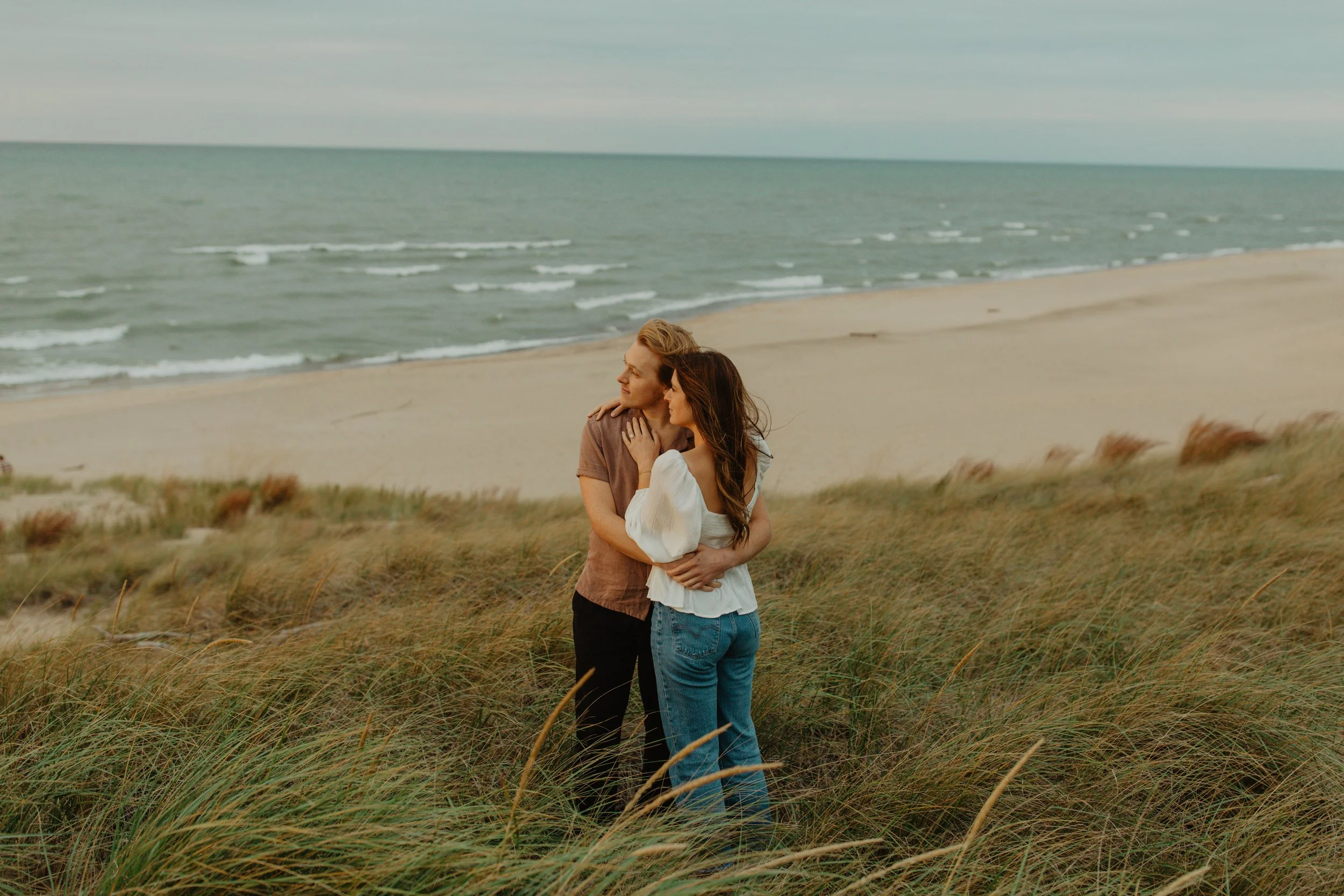 A young couple embracing on a grassy dune near the beach with ocean waves and cloudy sky in the background.
