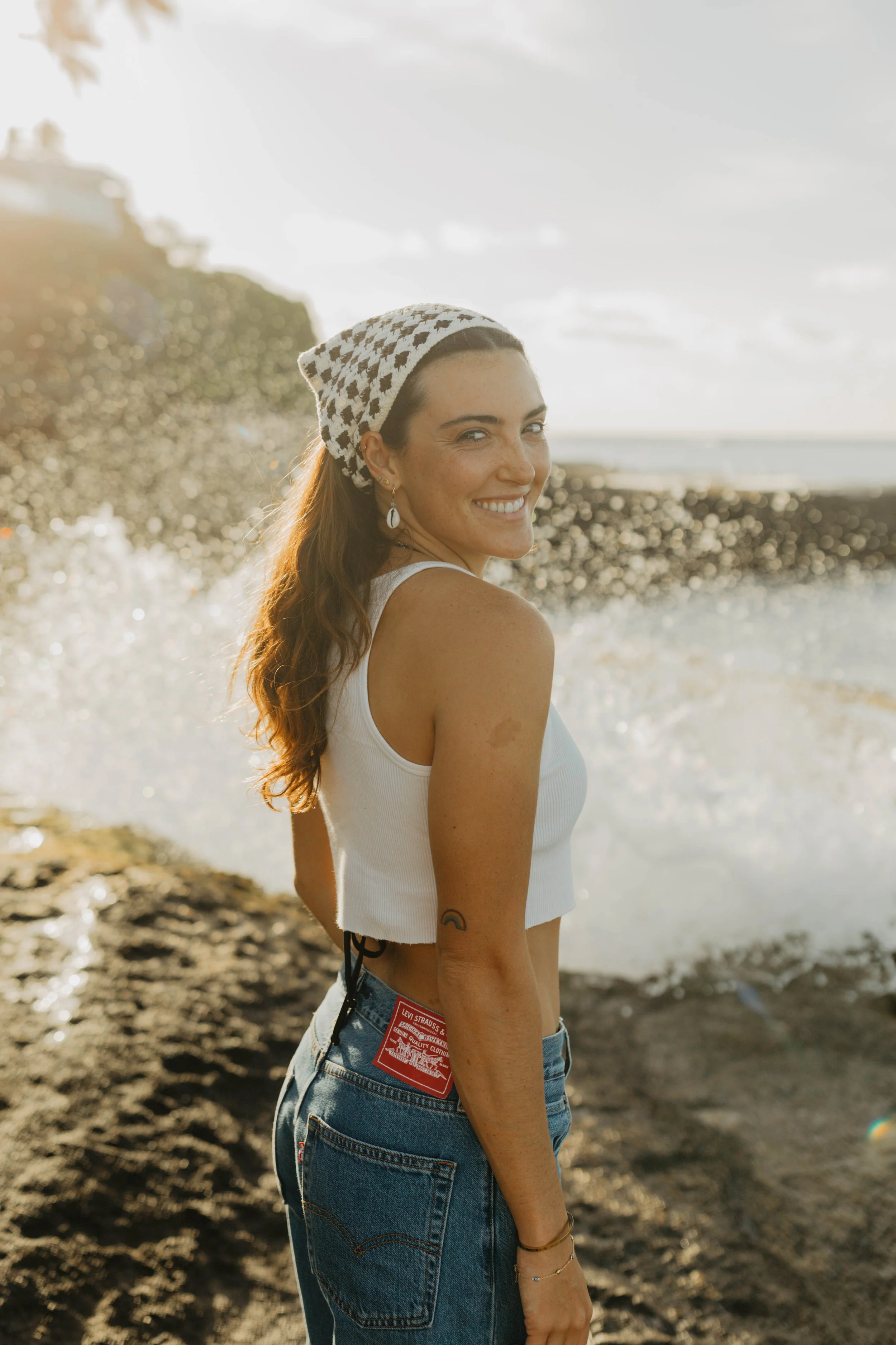 A woman smiling at the beach during sunset, wearing a white sleeveless top, denim jeans, earrings, and a patterned headscarf.