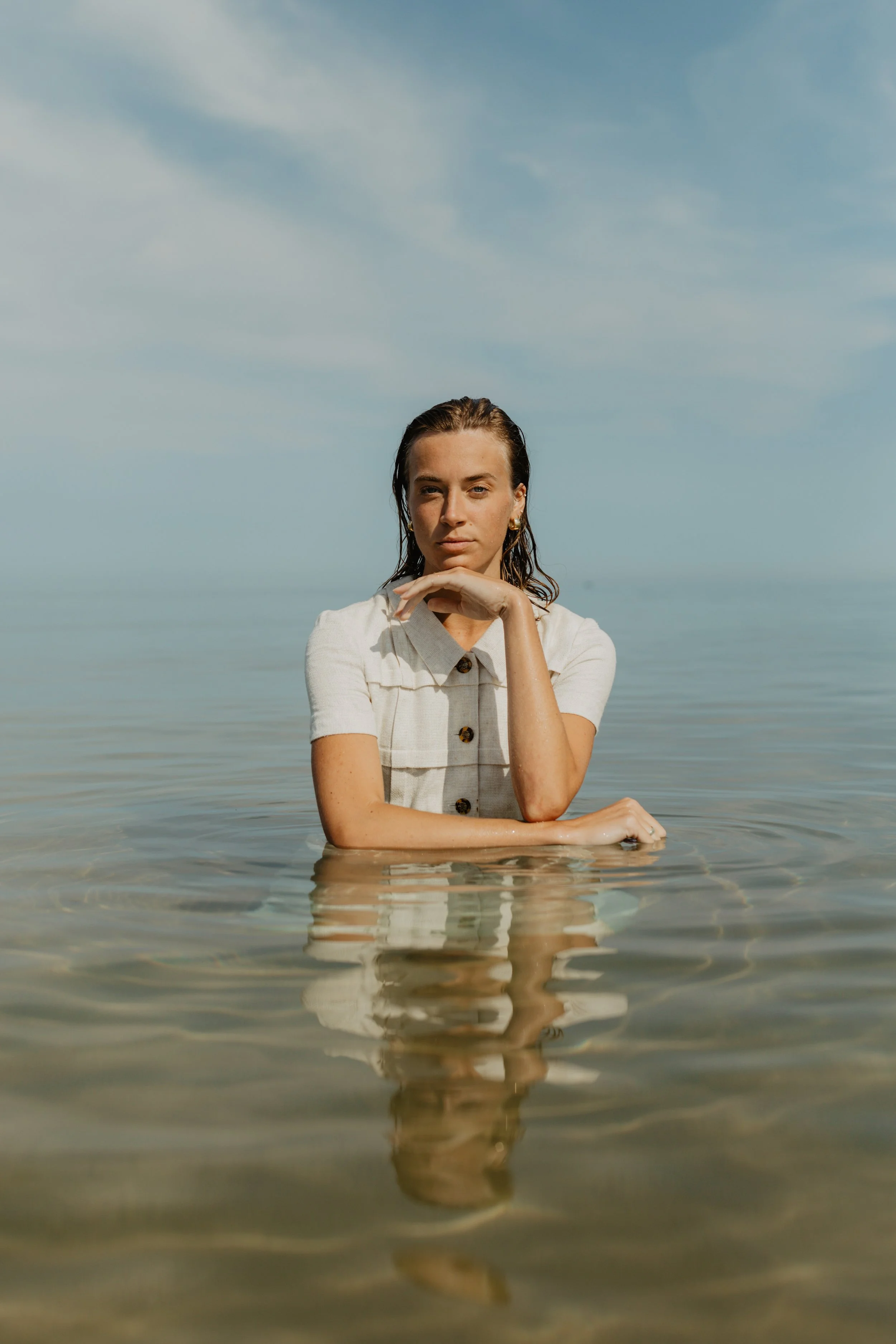 A woman standing in shallow water at the beach, with her chin resting on her hand, wearing a white short-sleeved shirt, and looking at the camera under blue sky.
