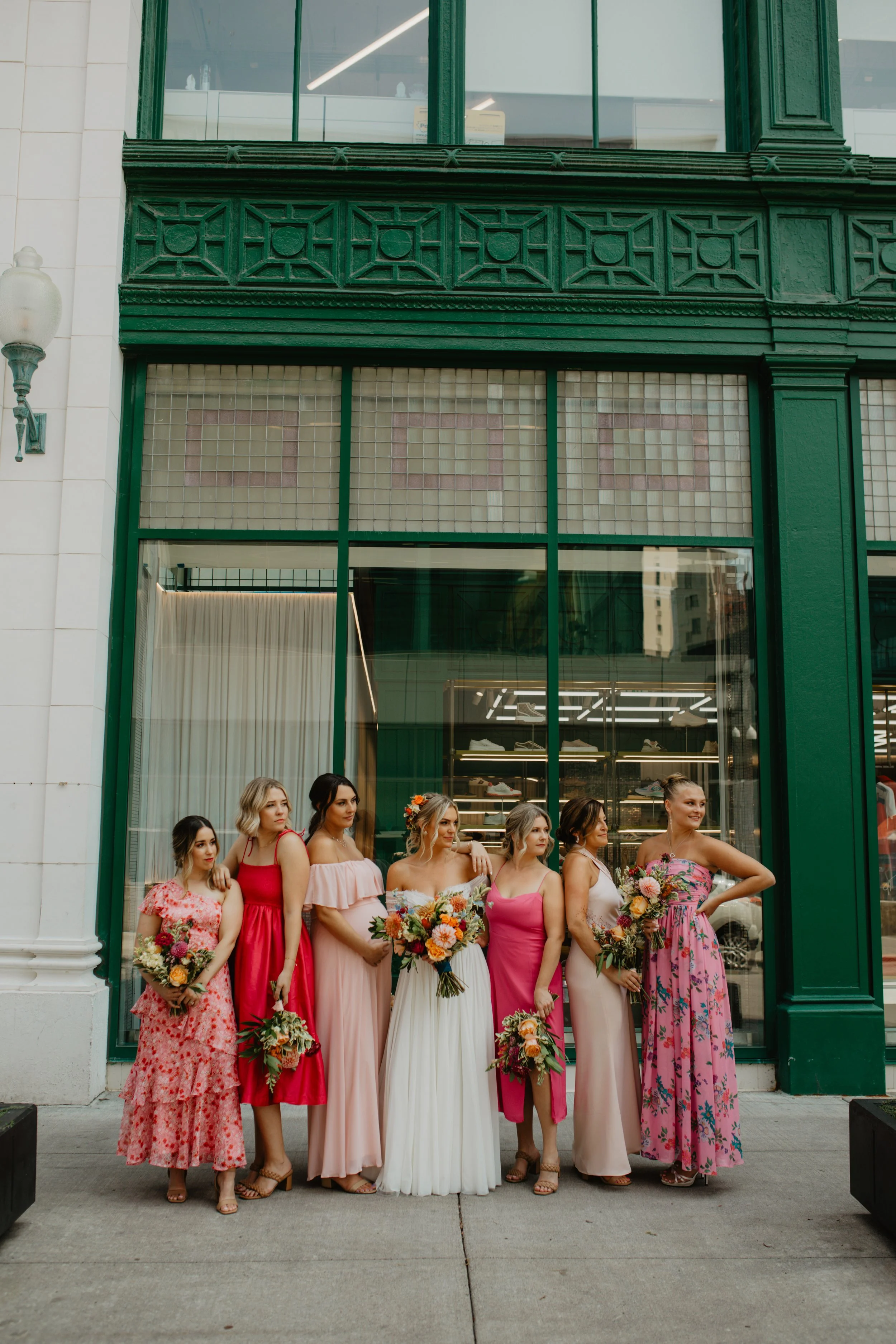A group of seven women dressed in colorful dresses, holding bouquets, standing outside a building with large glass windows and green decorative framing.