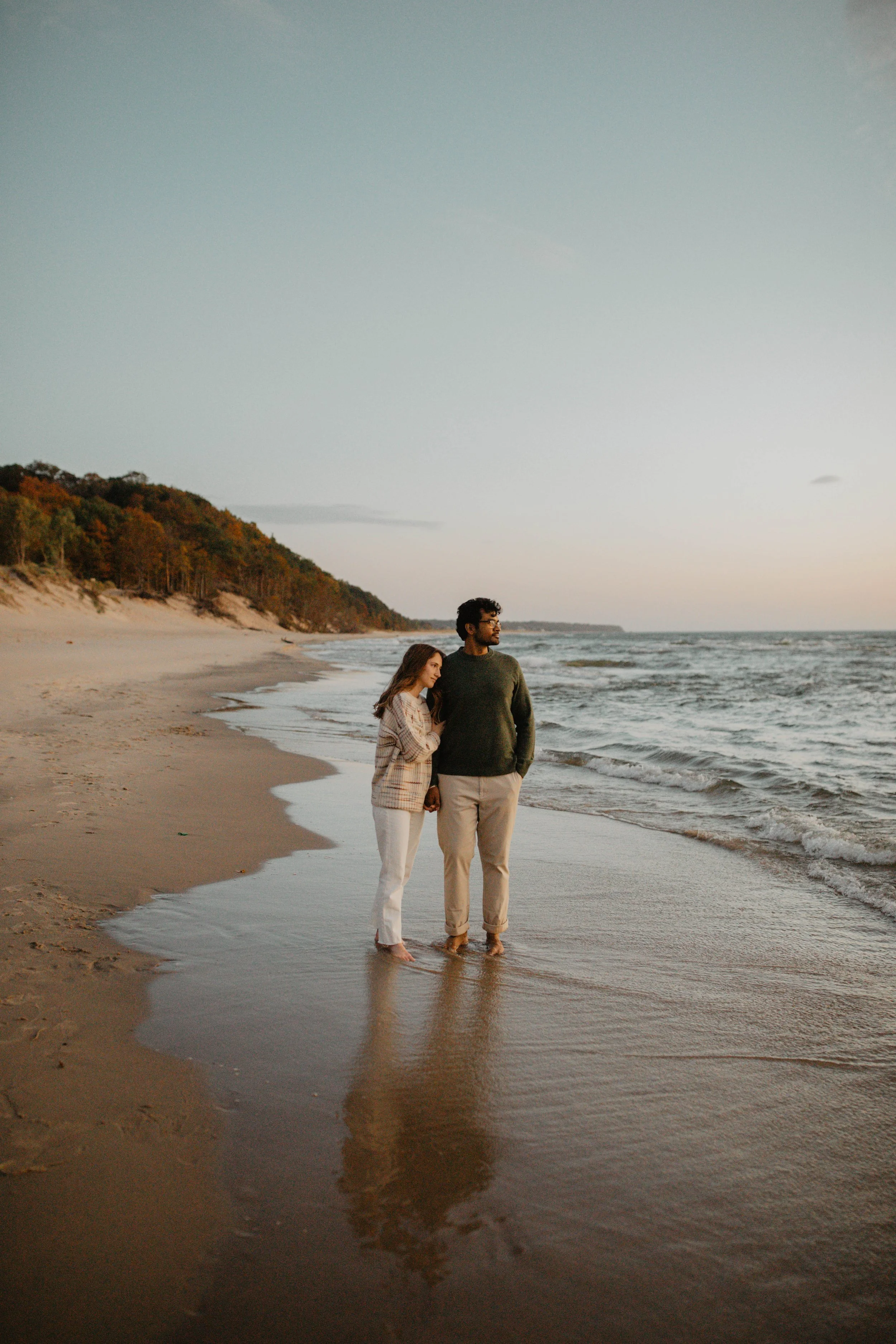 A couple standing barefoot on the sandy shoreline of a beach at sunset or dusk, holding hands and gazing towards the horizon.
