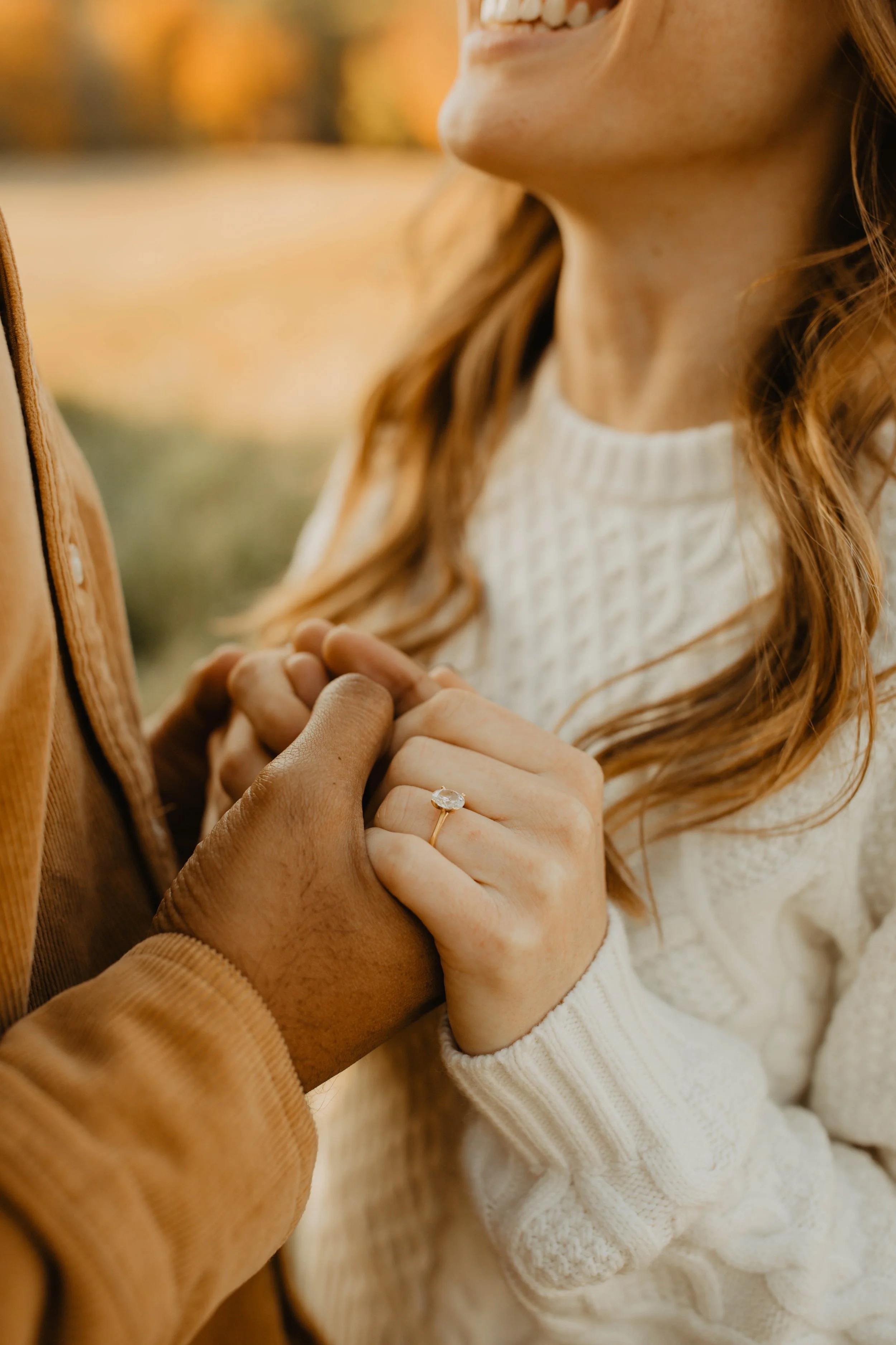 A woman and man hold hands, with the woman's hand displaying an engagement ring, outdoors during autumn.