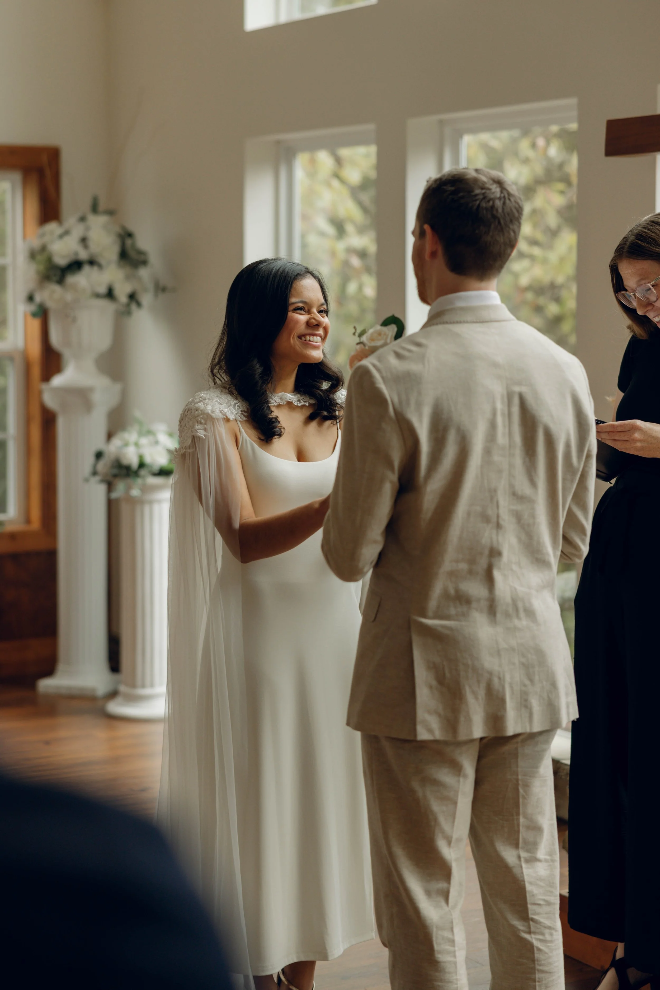A couple exchanging vows during a wedding ceremony in a bright room with large windows, with a woman officiant standing nearby.