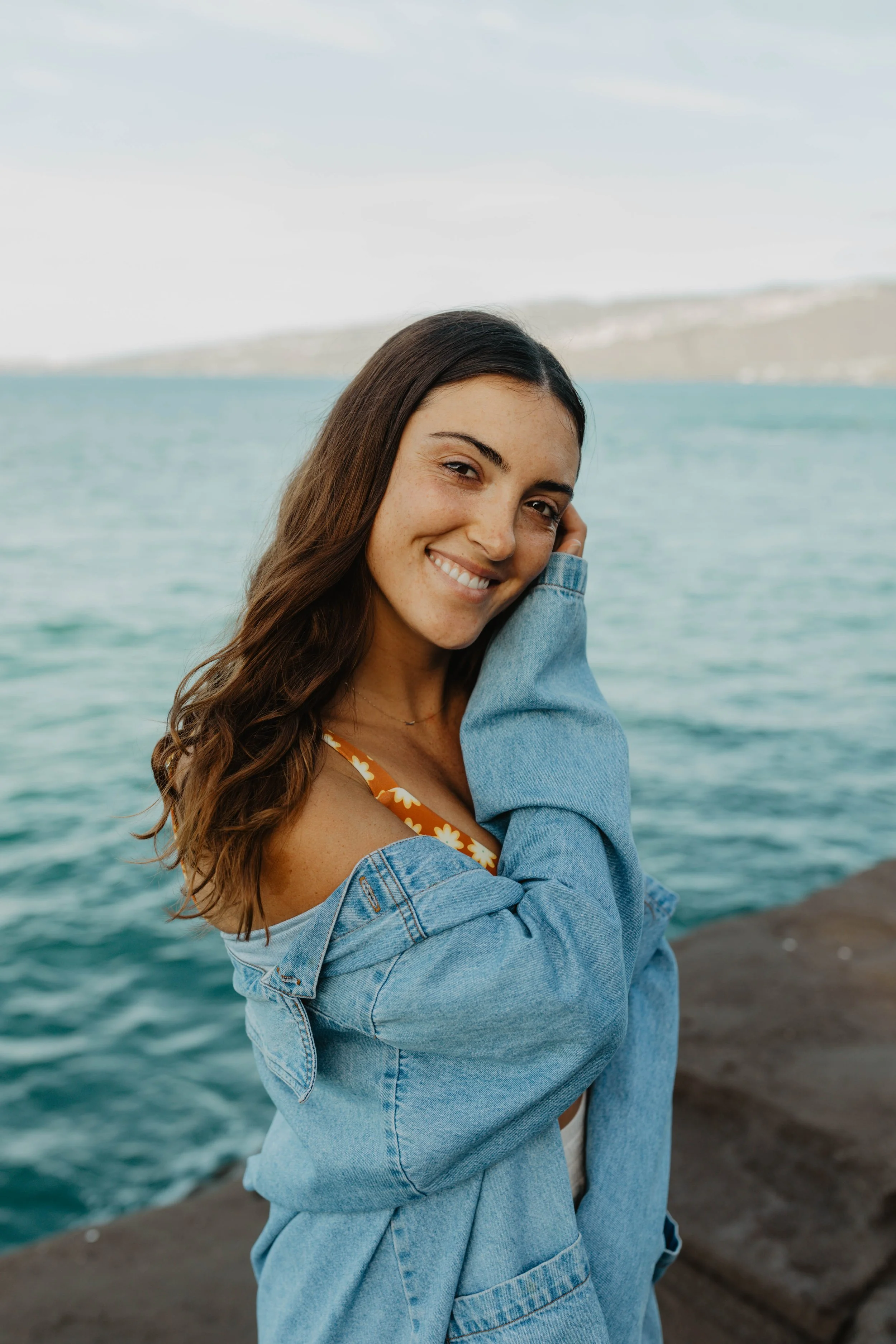 A young woman with long brown hair, smiling, wearing a denim jacket, standing near the water with a distant shoreline in the background.