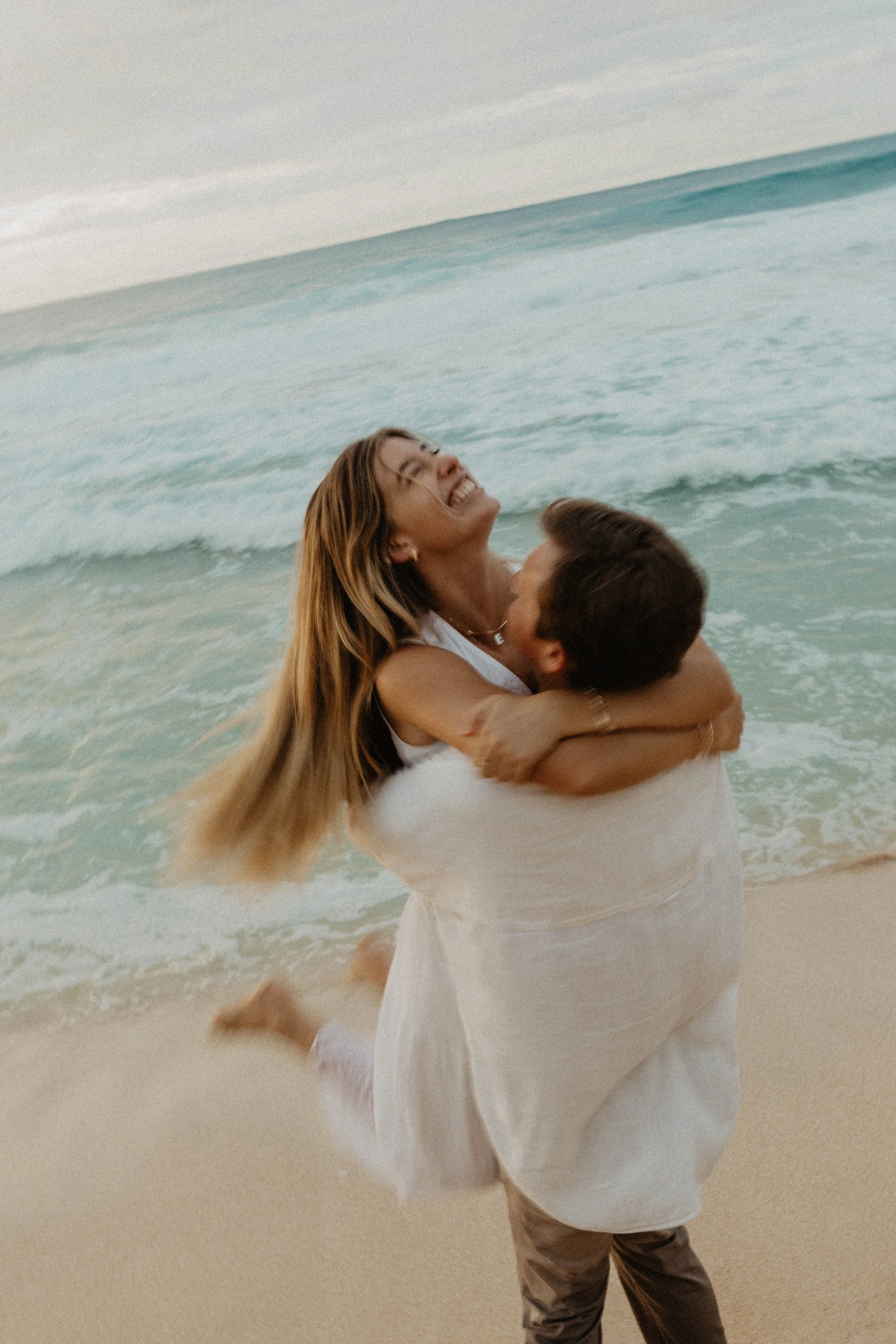 A woman in white dress being lifted by a man in beige pants on a sandy beach with ocean waves in the background, both smiling and enjoying the moment.