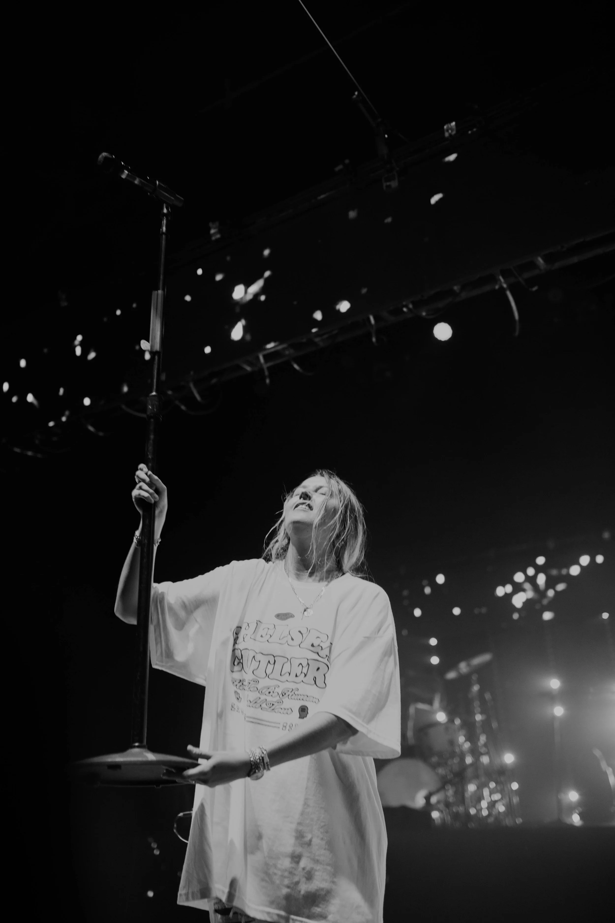 Chelsea Cutler sings on stage holding a microphone stand with one hand, wearing an oversized t-shirt, performing with her eyes closed in a dark concert setting.