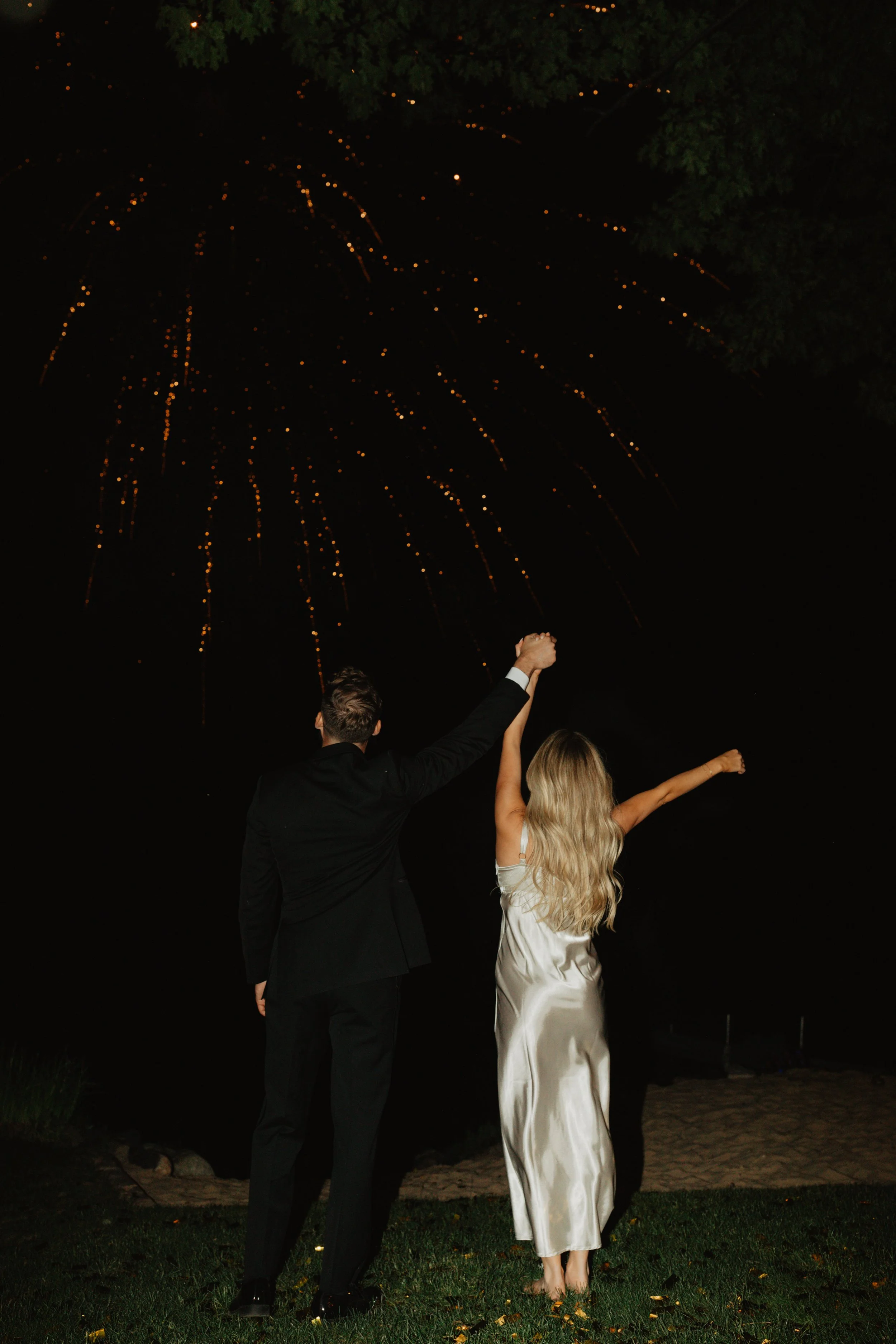A couple celebrating outdoors at night, holding hands up with fireworks in the sky above them.