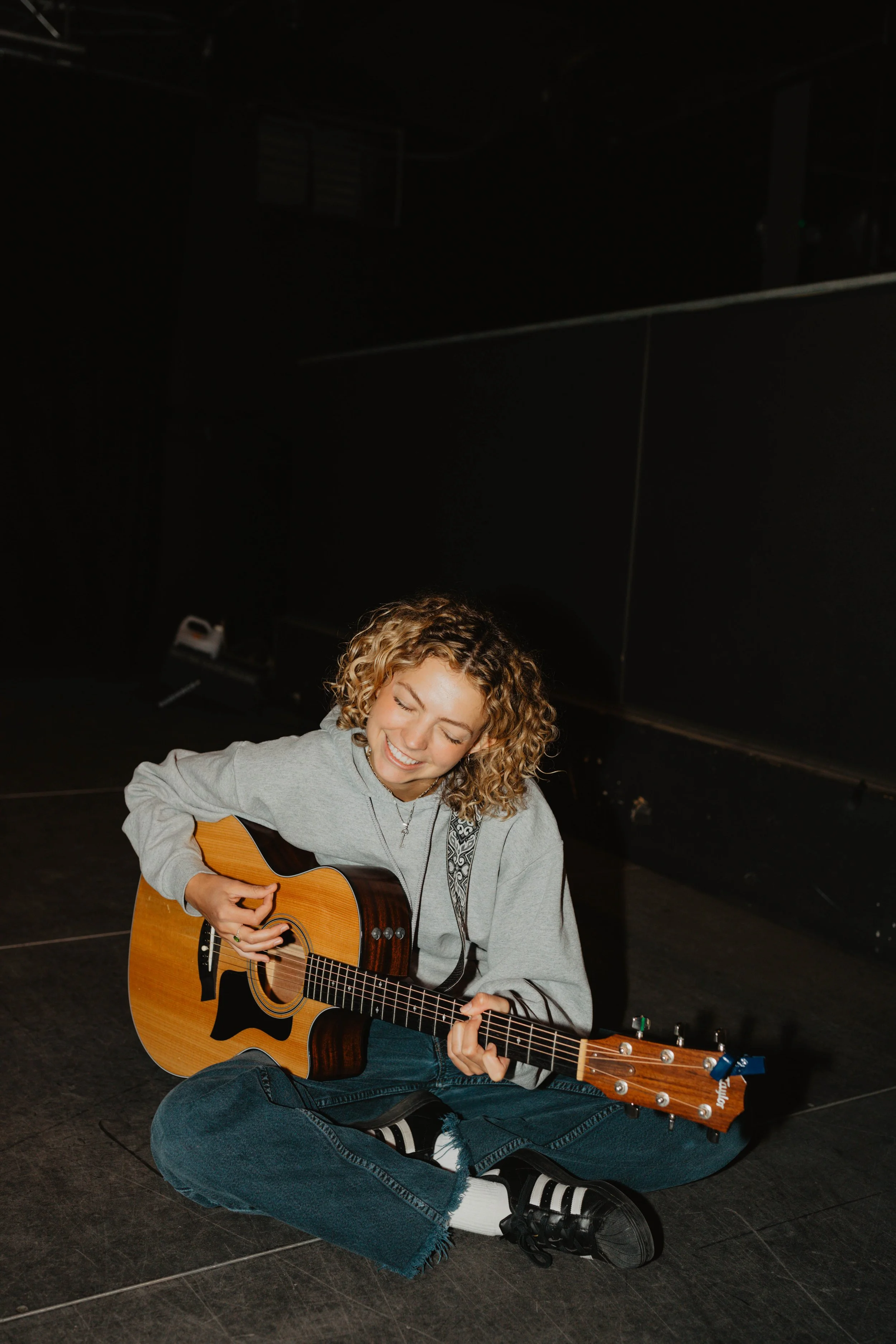 Young woman sitting on the floor playing an acoustic guitar, smiling with eyes closed, in a dark room.