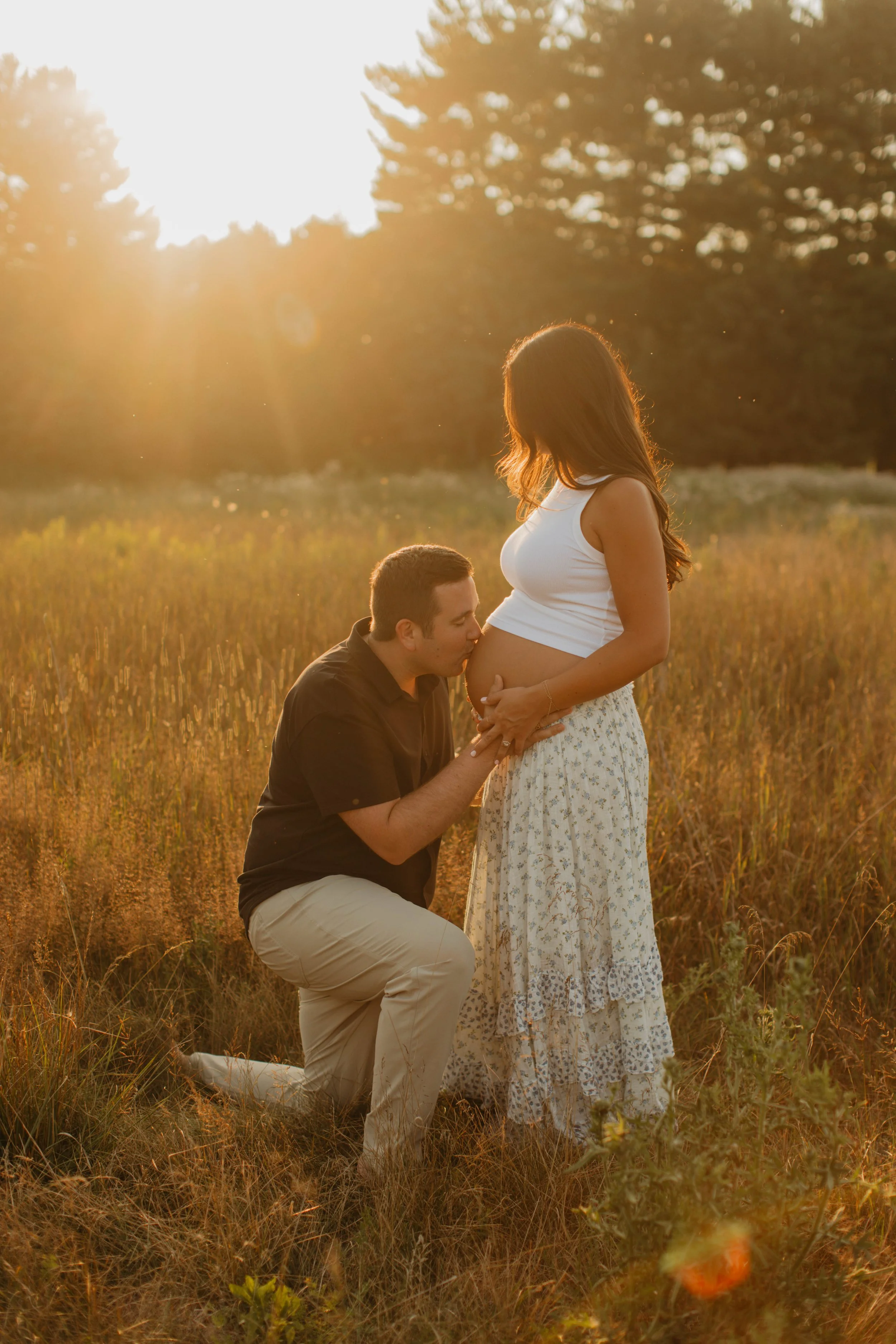 A man kneeling and kissing a pregnant woman's belly during sunset in a grassy field.