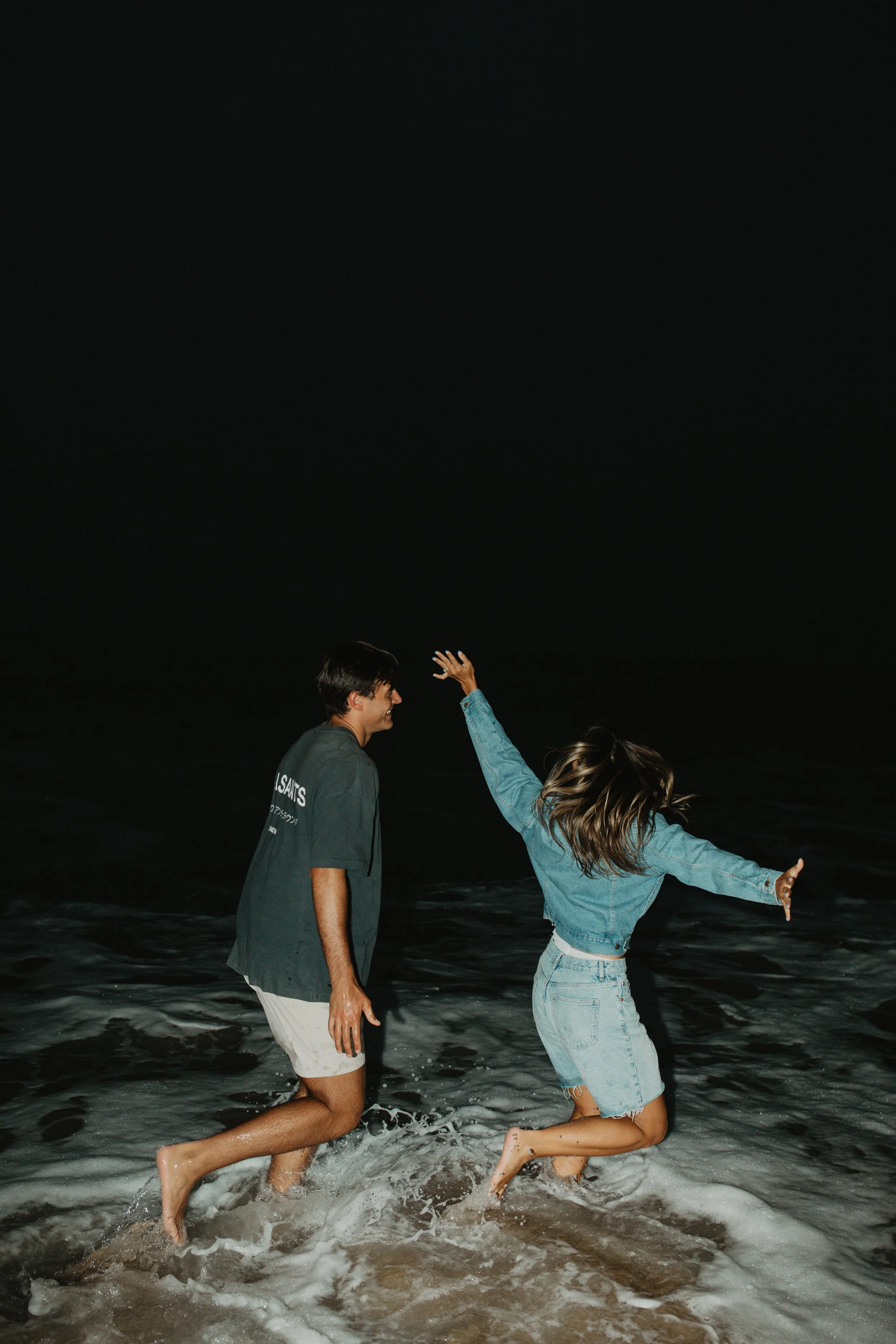 Two people, a man and a woman, playing and jumping in the ocean at night.