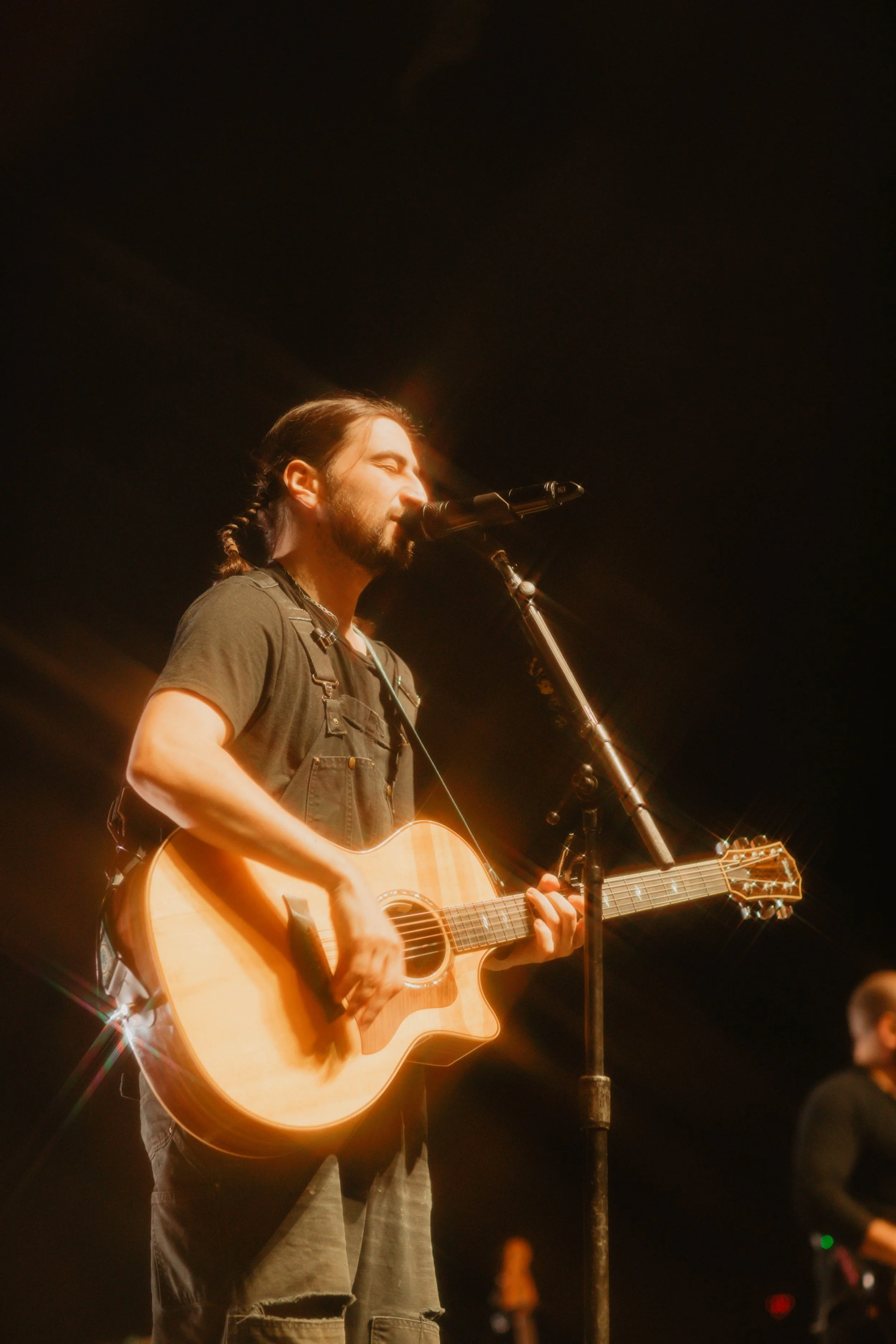 Noah Kahanplaying an acoustic guitar and singing into a microphone on stage under warm lighting in Detroit, Michigan.
