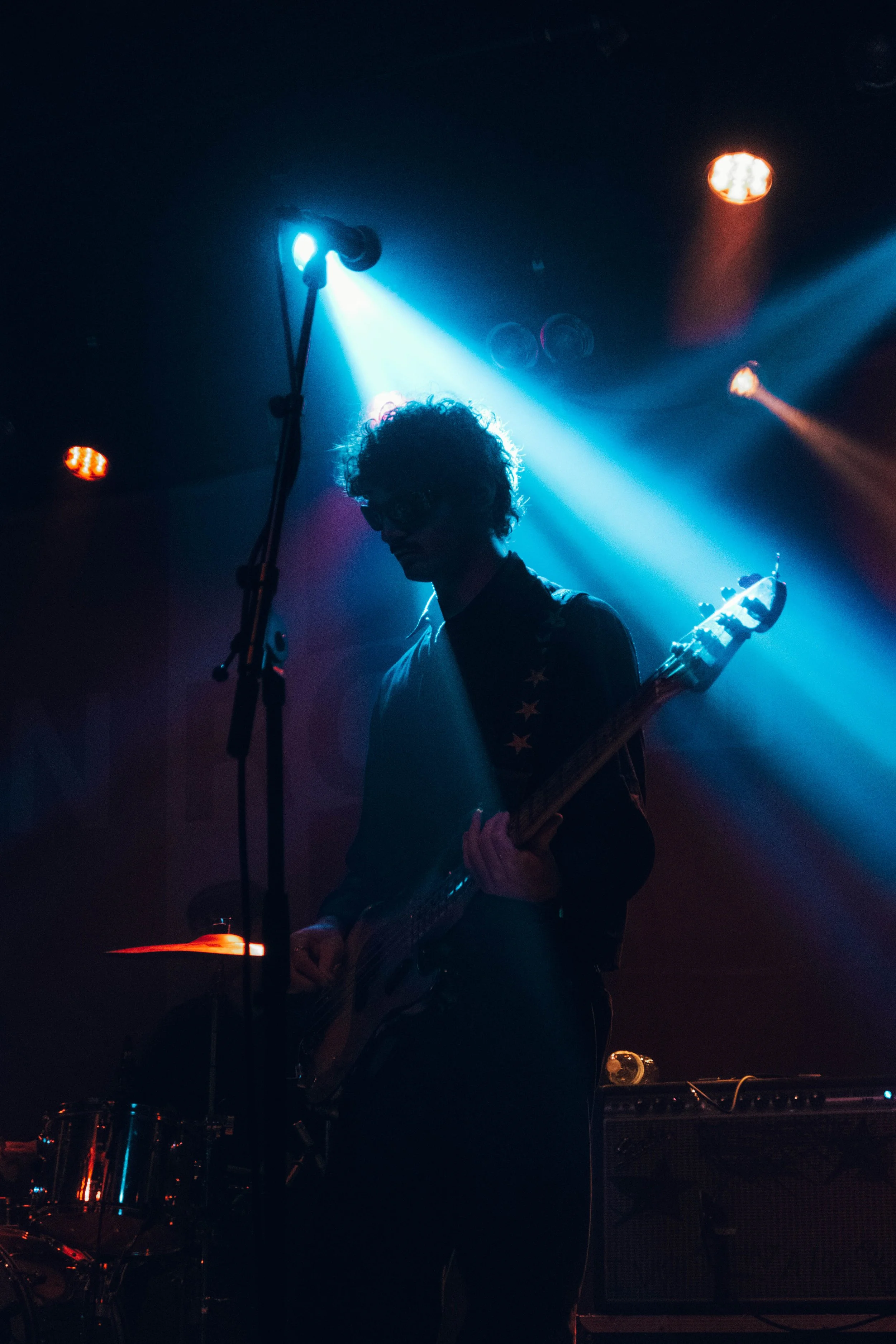 The band SUNROOM playing an electric guitar on stage, illuminated by blue and orange stage lights, with a microphone stand in the foreground at their concert in Chicago.