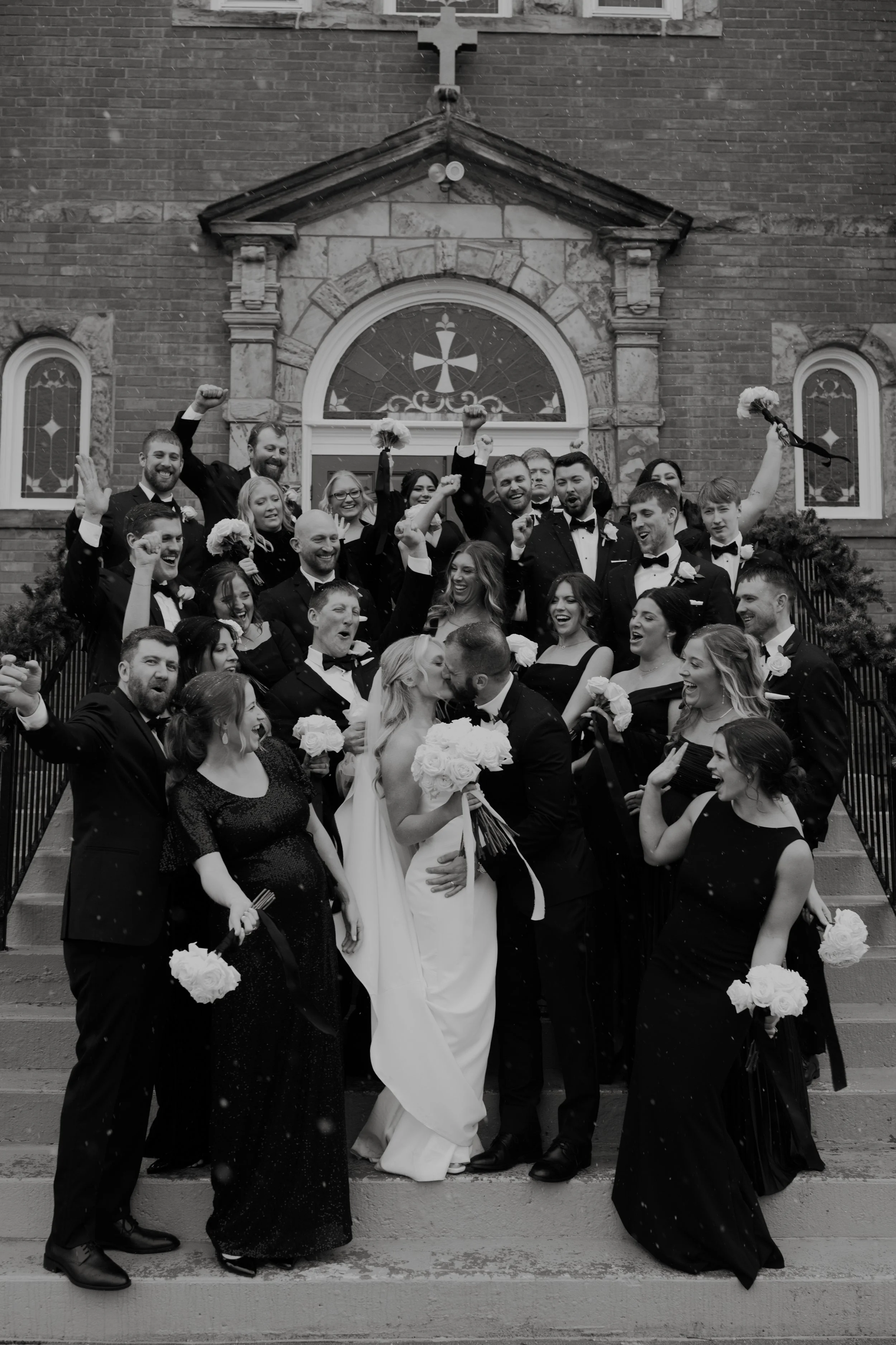 A group of wedding guests and the bride and groom on church steps, celebrating and posing for a photo. The bride and groom are kissing in the center, holding a bouquet of flowers, surrounded by guests in formal attire.