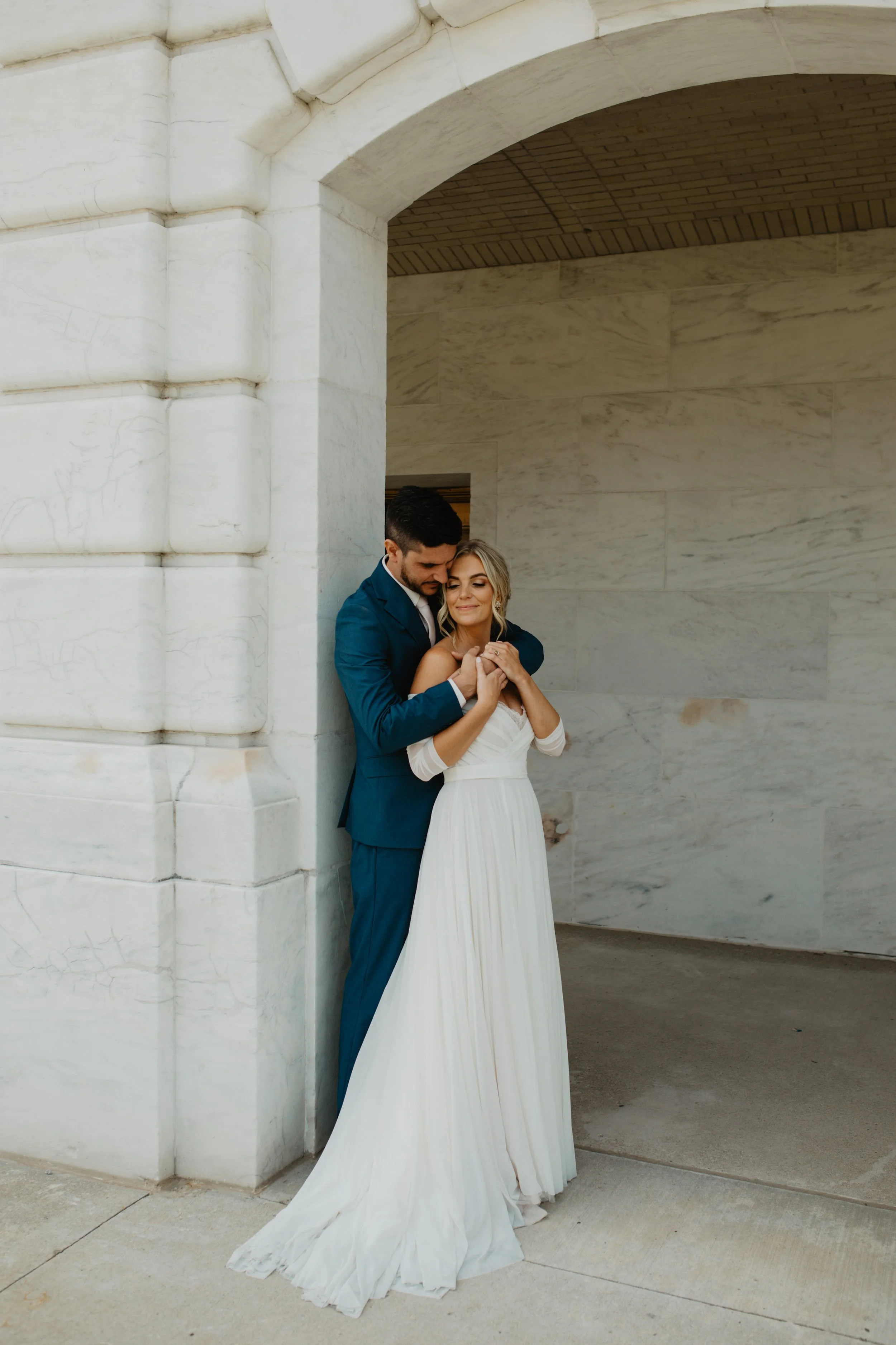A bride and groom embrace under an archway with marble walls, the groom in a blue suit and the bride in a flowing white dress.