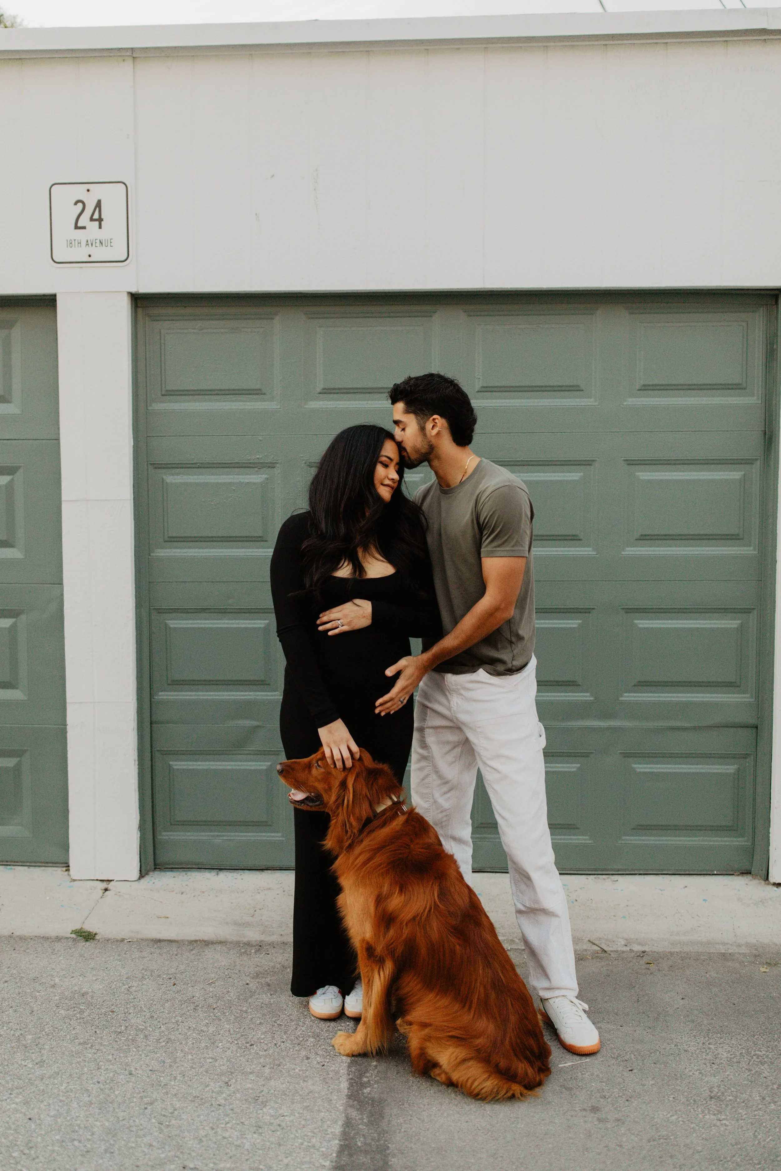 A man and woman standing outside in front of a green garage door, with the woman being pregnant, a dog sitting in front of them, woman dressed in black, man in gray t-shirt and white pants, on 18th Avenue.