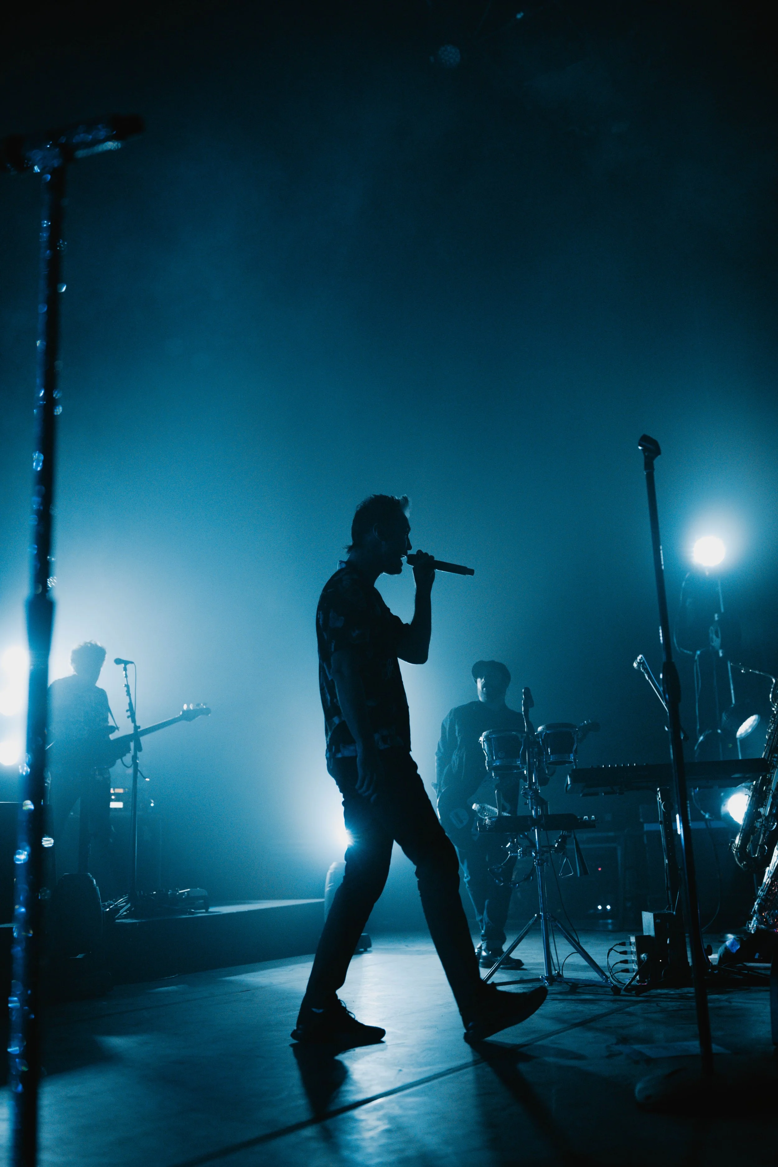 Silhouette photo of the band, Fitz & the Tantrums performing on stage with band members playing instruments in the background, illuminated by blue stage lights.