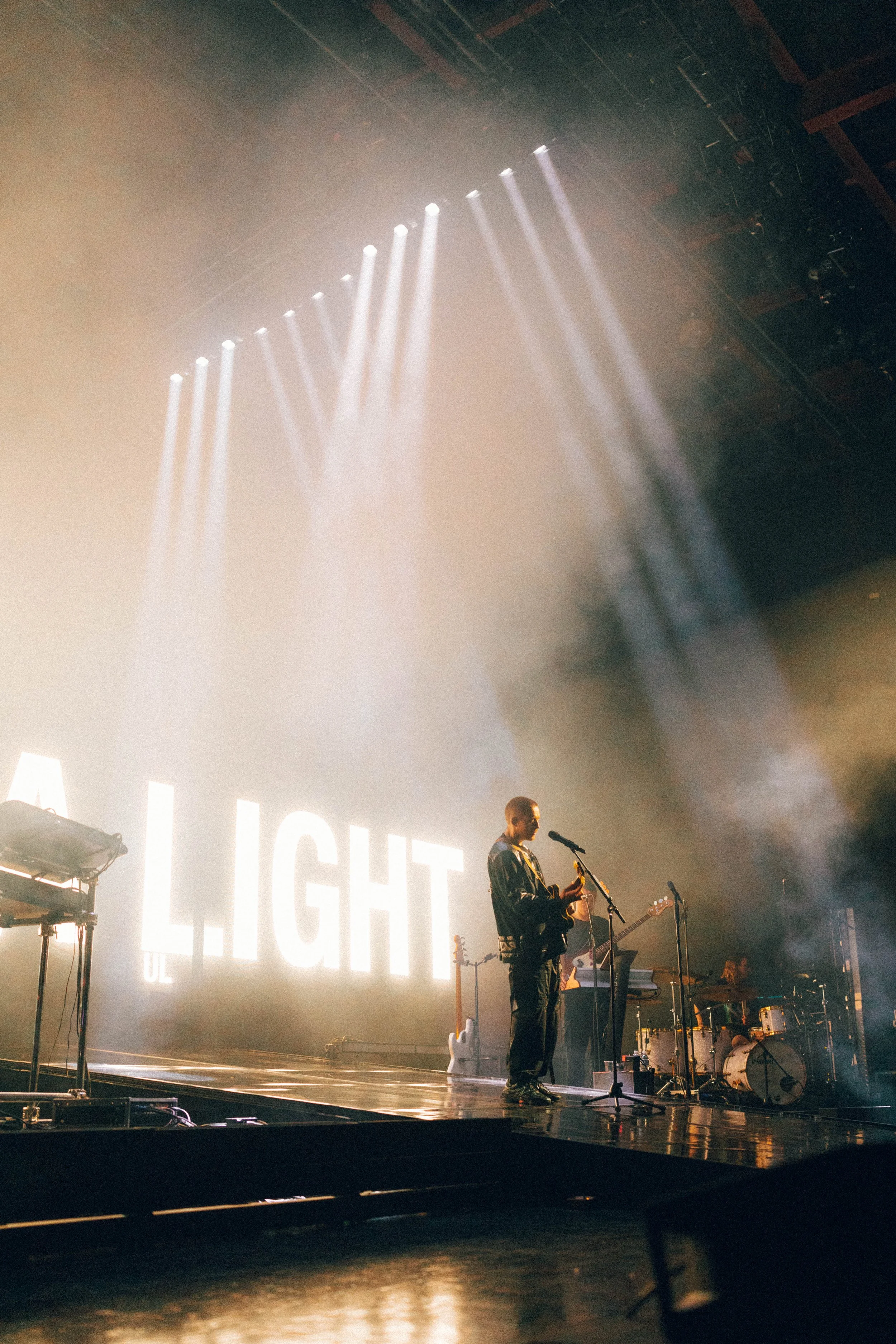 Dermot Kennedy performs on a stage with bright lighting in Detroit, Michigan. A musician is playing an electric guitar in front of a large sign that says 'LIGHT'.