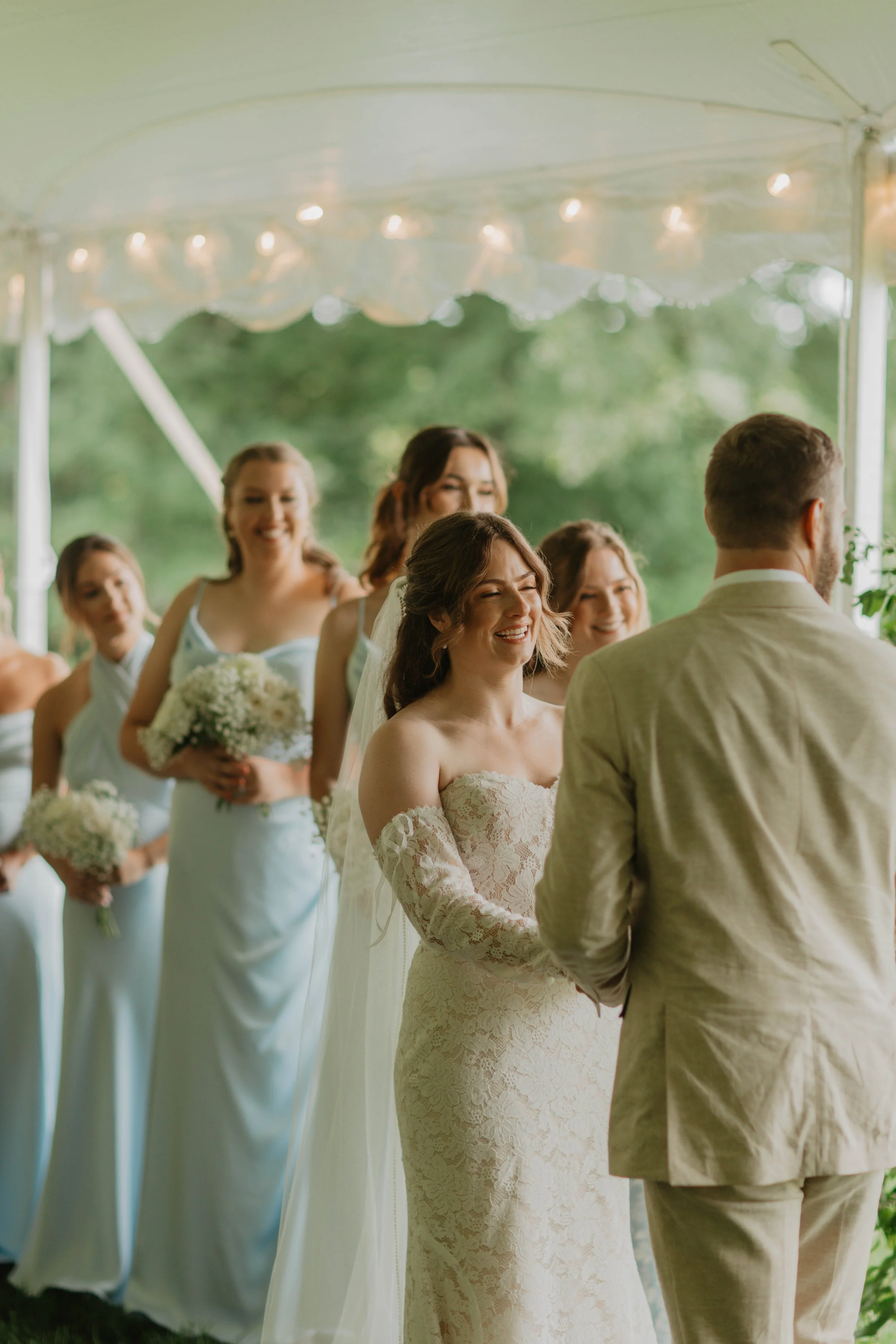 A bride and groom exchanging vows at an outdoor wedding under a white tent, with bridesmaids holding bouquets in the background.