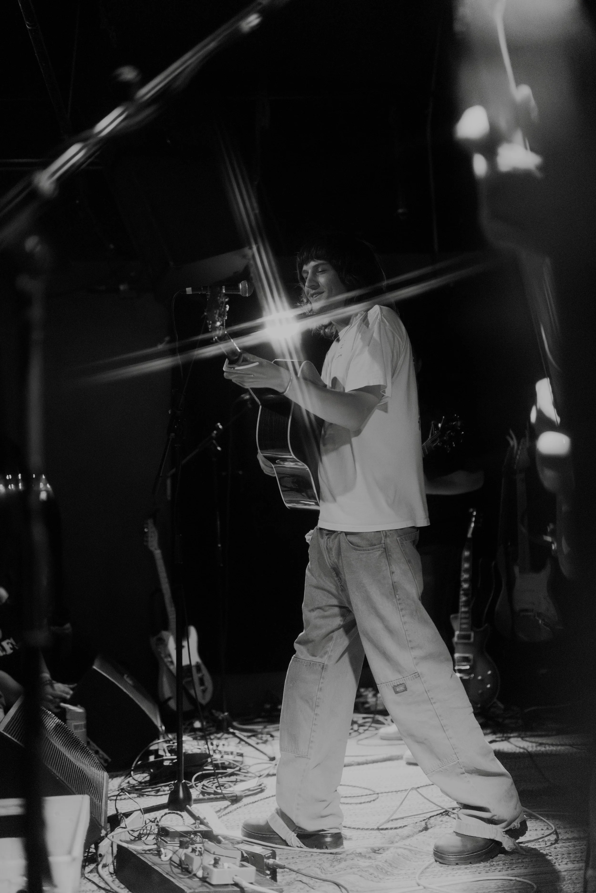 The Band Dipsea Flower plays  an acoustic guitar, singing, and smiling on stage in a dimly lit setting, with musical instruments and equipment visible around.