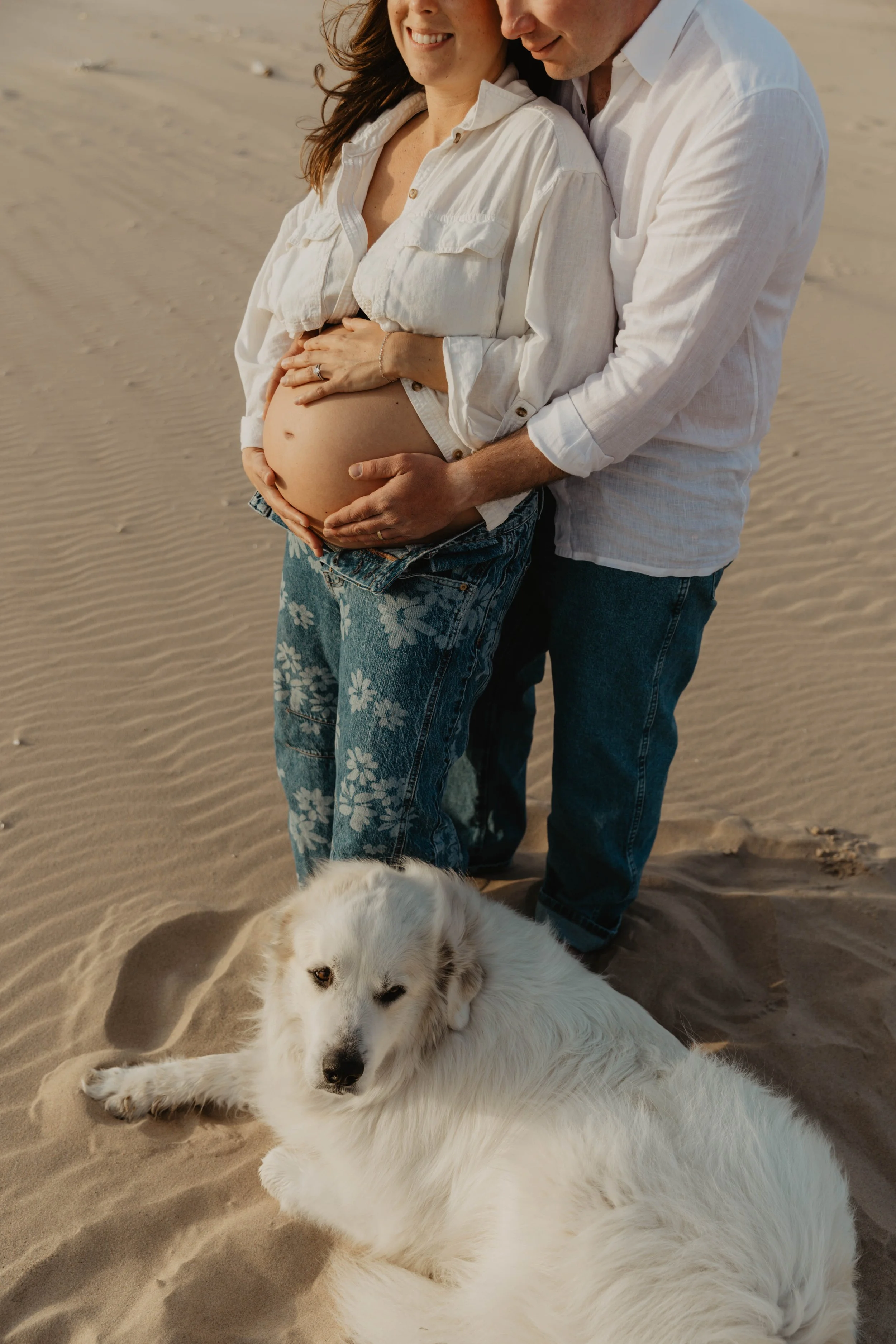 A pregnant woman and a man on the beach, with the man holding her baby bump, and a white dog lying on the sand in front of them.