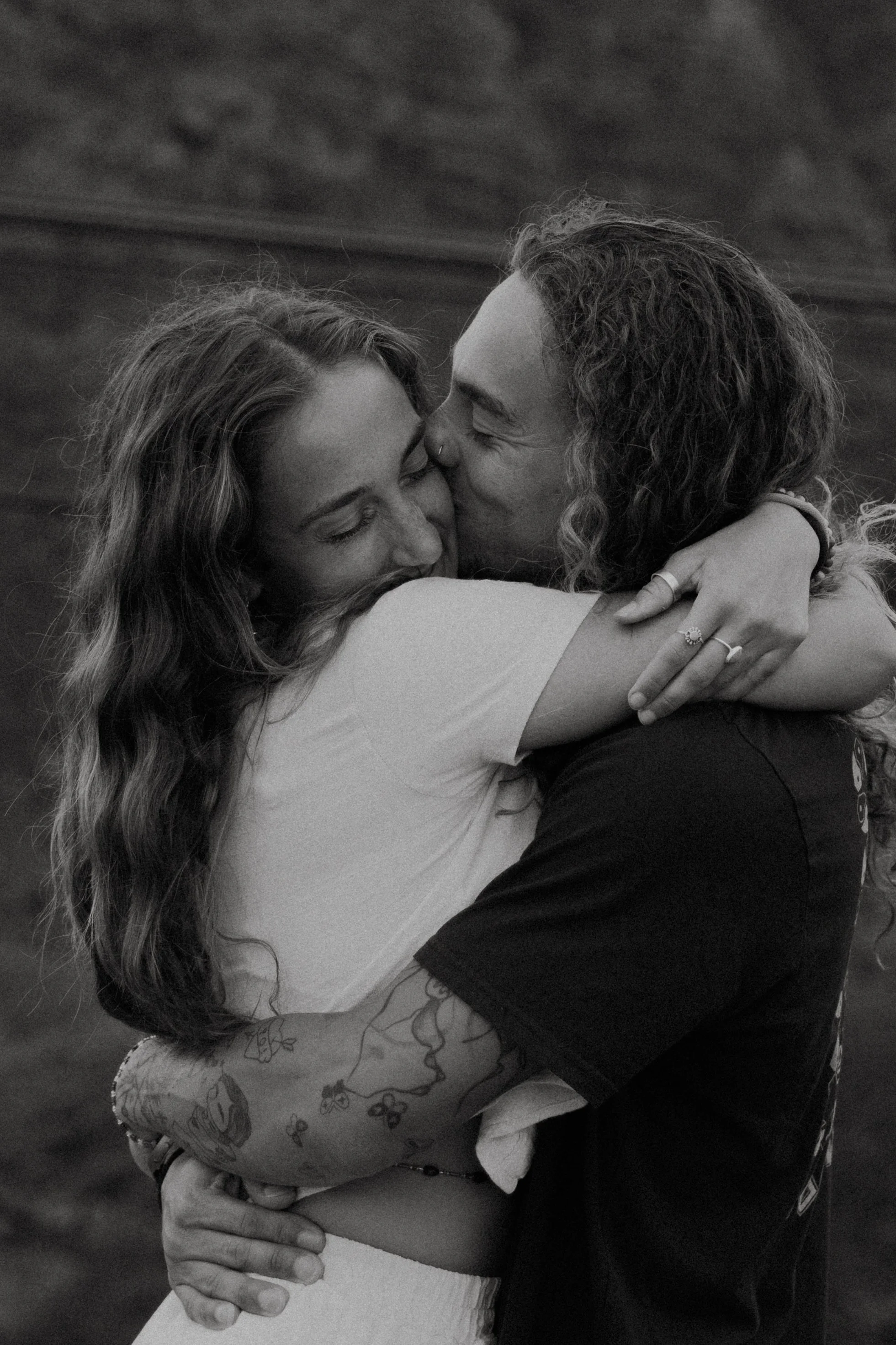 A black and white photo of a couple hugging and kissing outdoors, with a blurred landscape in the background.