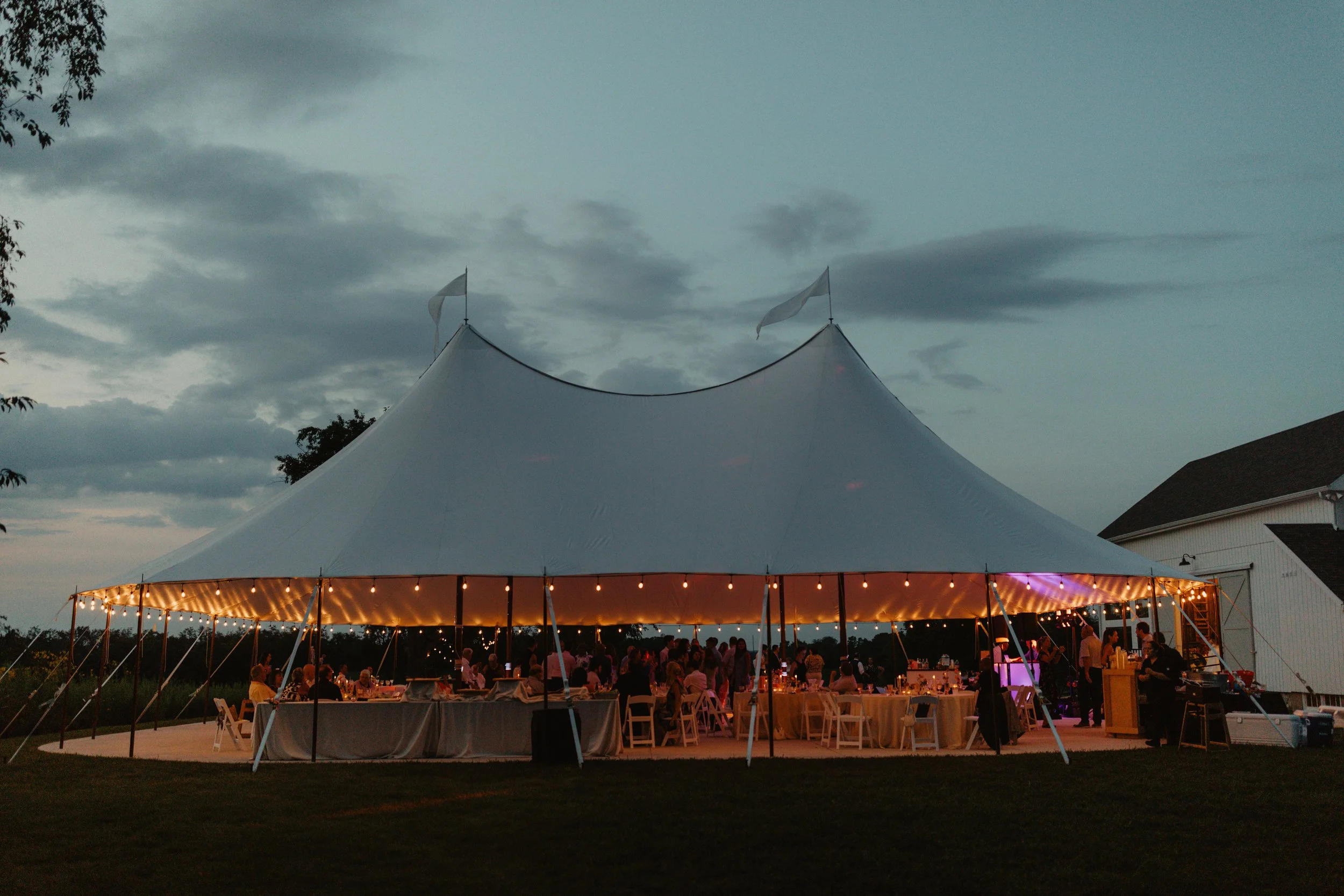 Large white event tent with string lights at dusk, with people gathered inside and around tables, adjacent to a building on the right, under a cloudy sky.