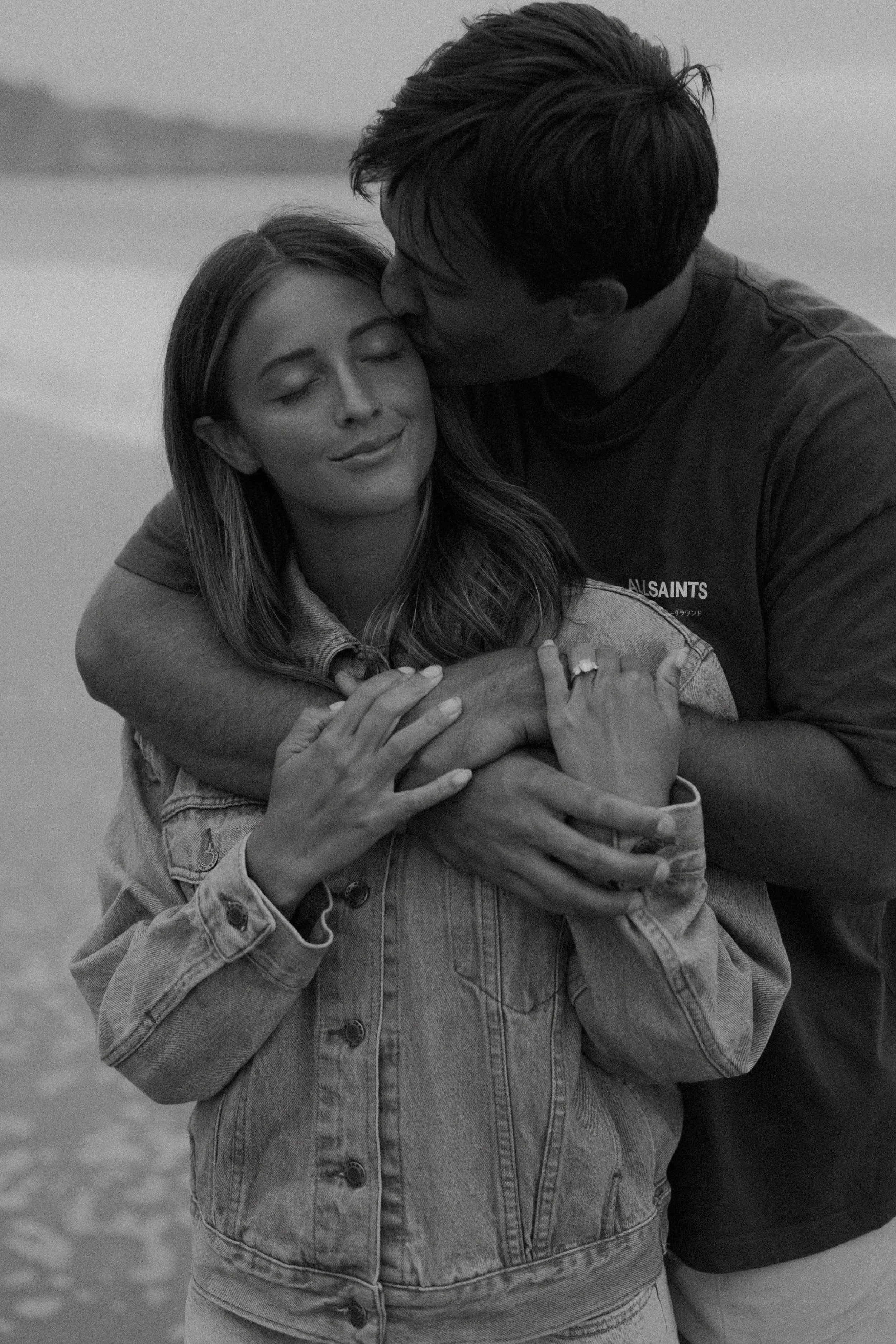 A black-and-white photo of a man kissing a woman on the forehead, both with their eyes closed, embracing on a beach.