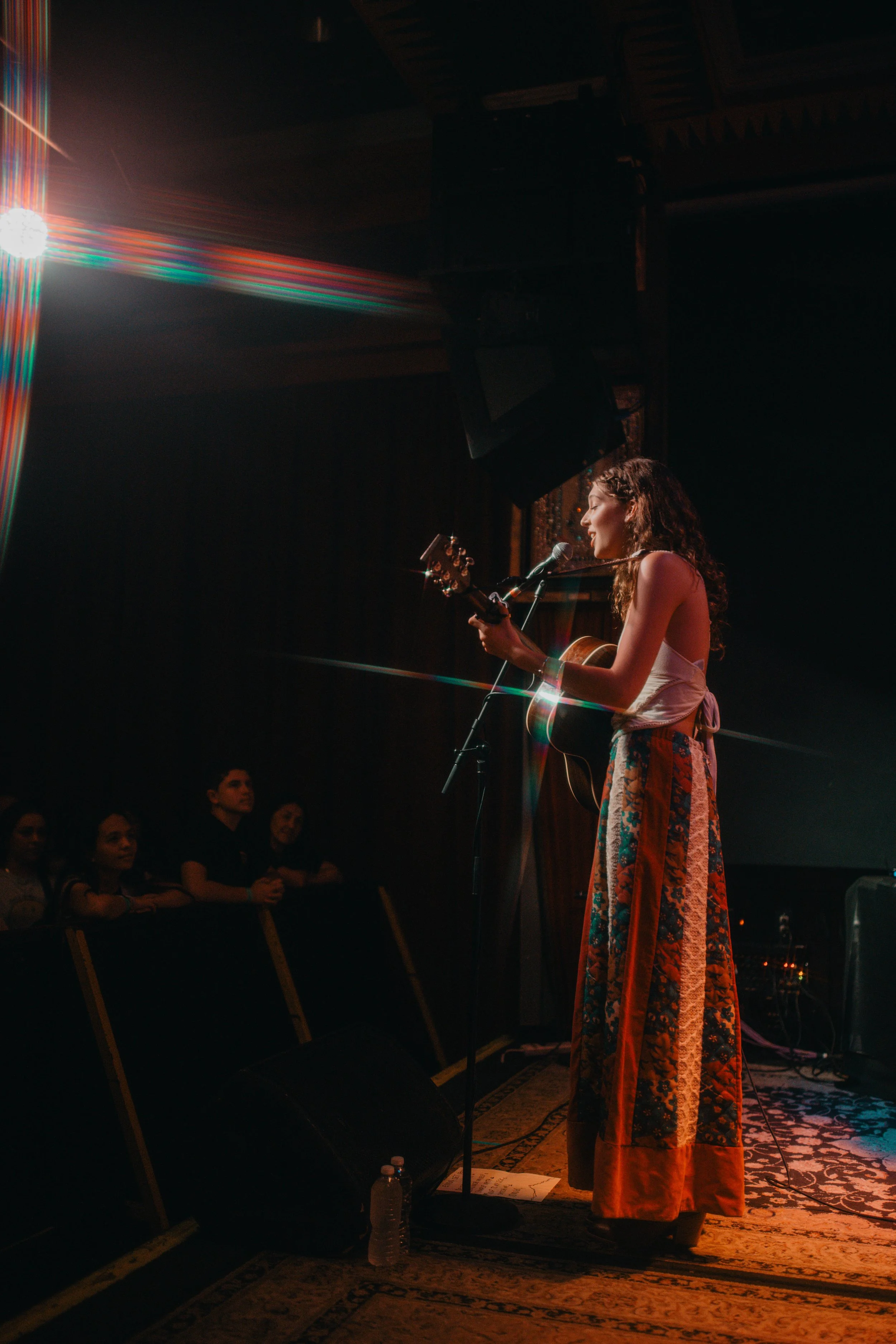 Emma Andersen, a  singer performing on stage with guitar, microphone, and colorful stage lighting, while audience watches attentively in a dark concert venue.