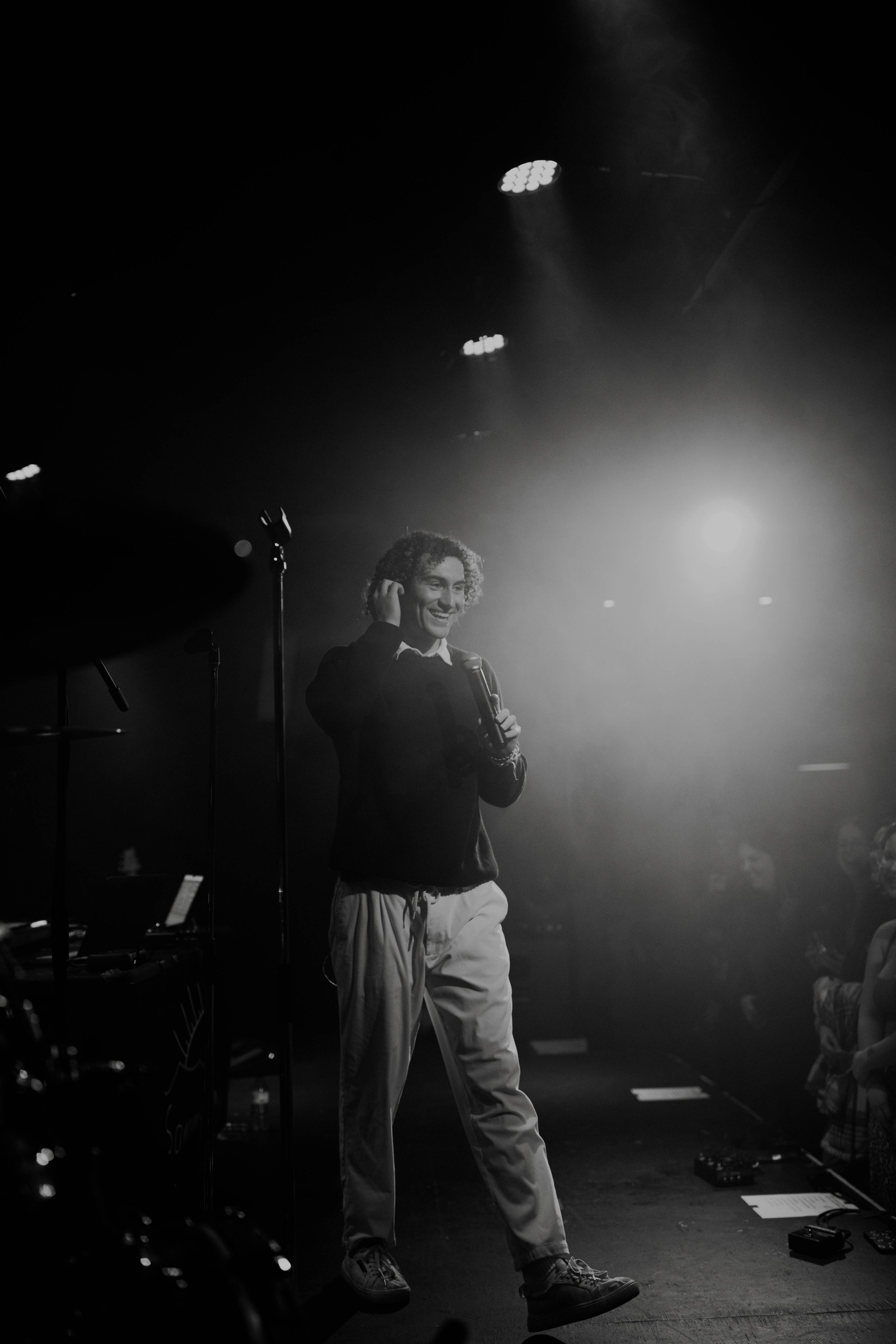 Sammy Rash on a stage, smiling, holding a microphone, under bright stage lights, with an audience visible in the background.