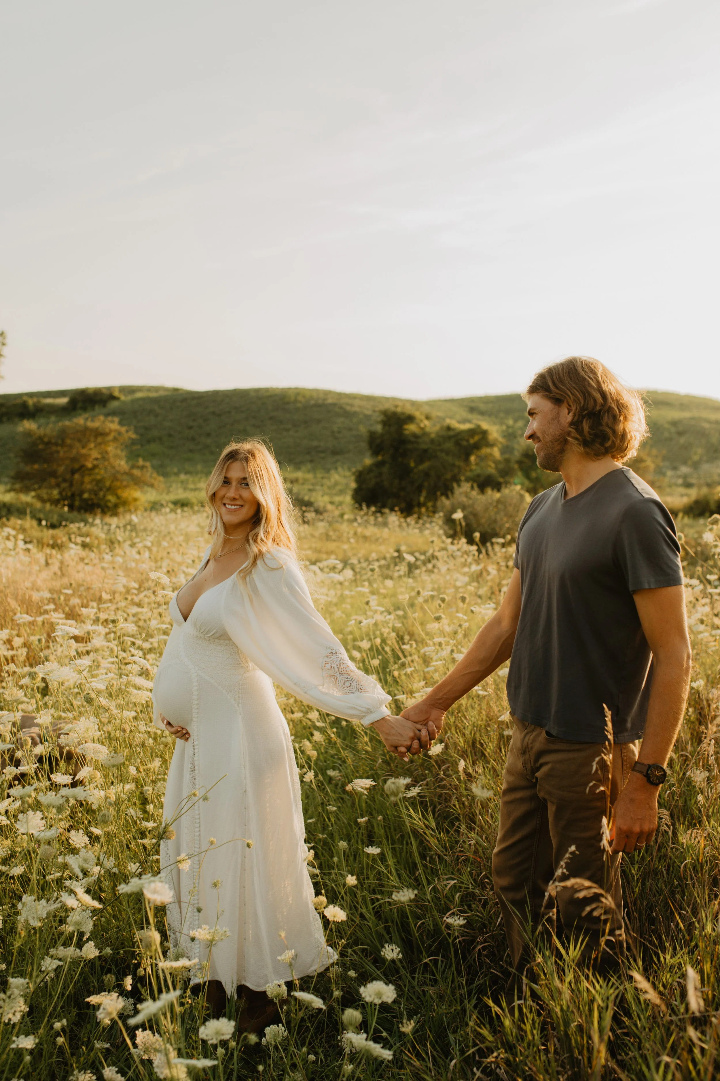 Pregnant woman in white dress holding hands with man in black T-shirt in a field of wildflowers during sunset.
