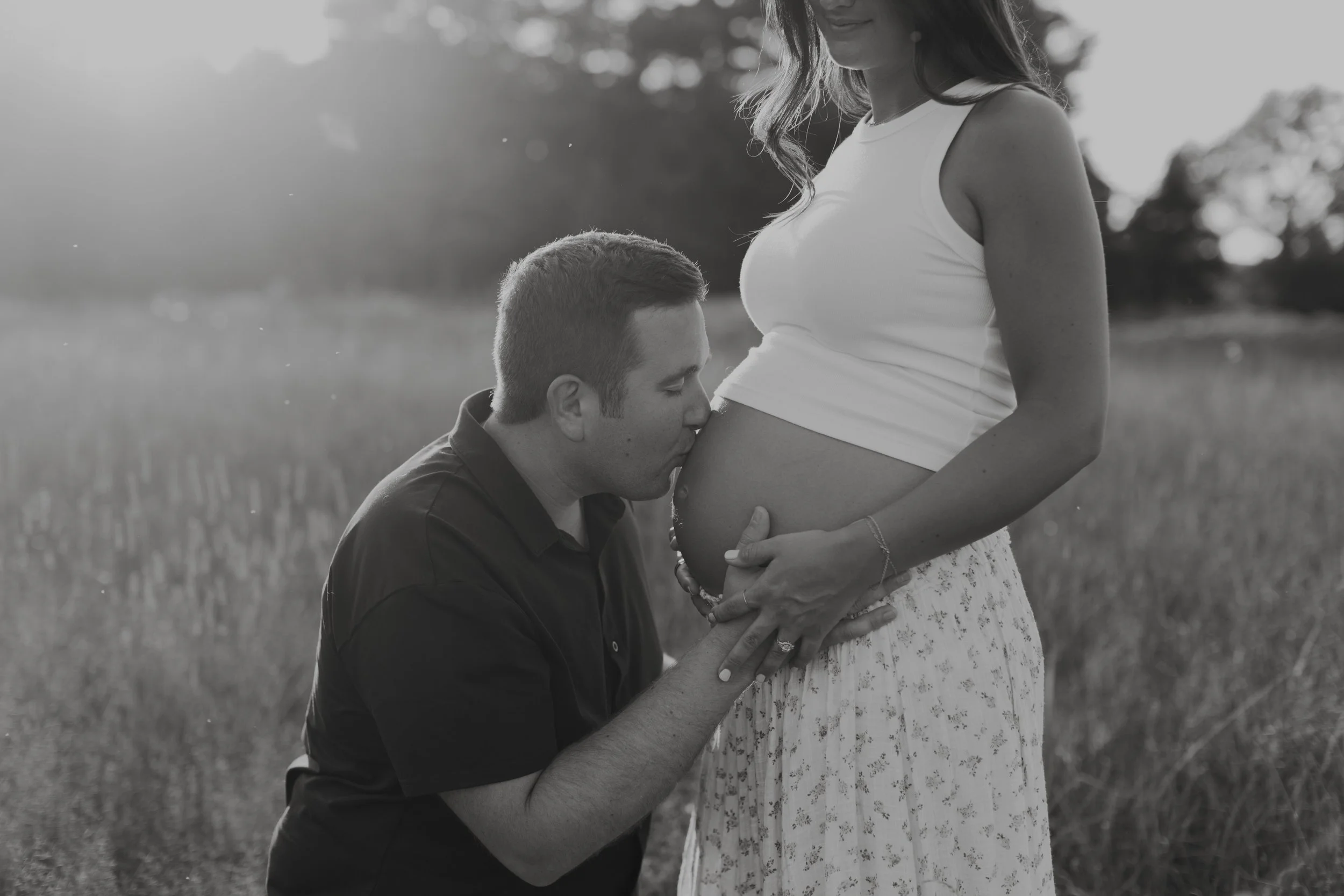 A man kissing a pregnant woman on her belly outdoors in a grassy field. The woman is standing, wearing a white tank top and a long skirt, and holding her belly. The man is kneeling, leaning in to kiss her belly with his eyes closed.