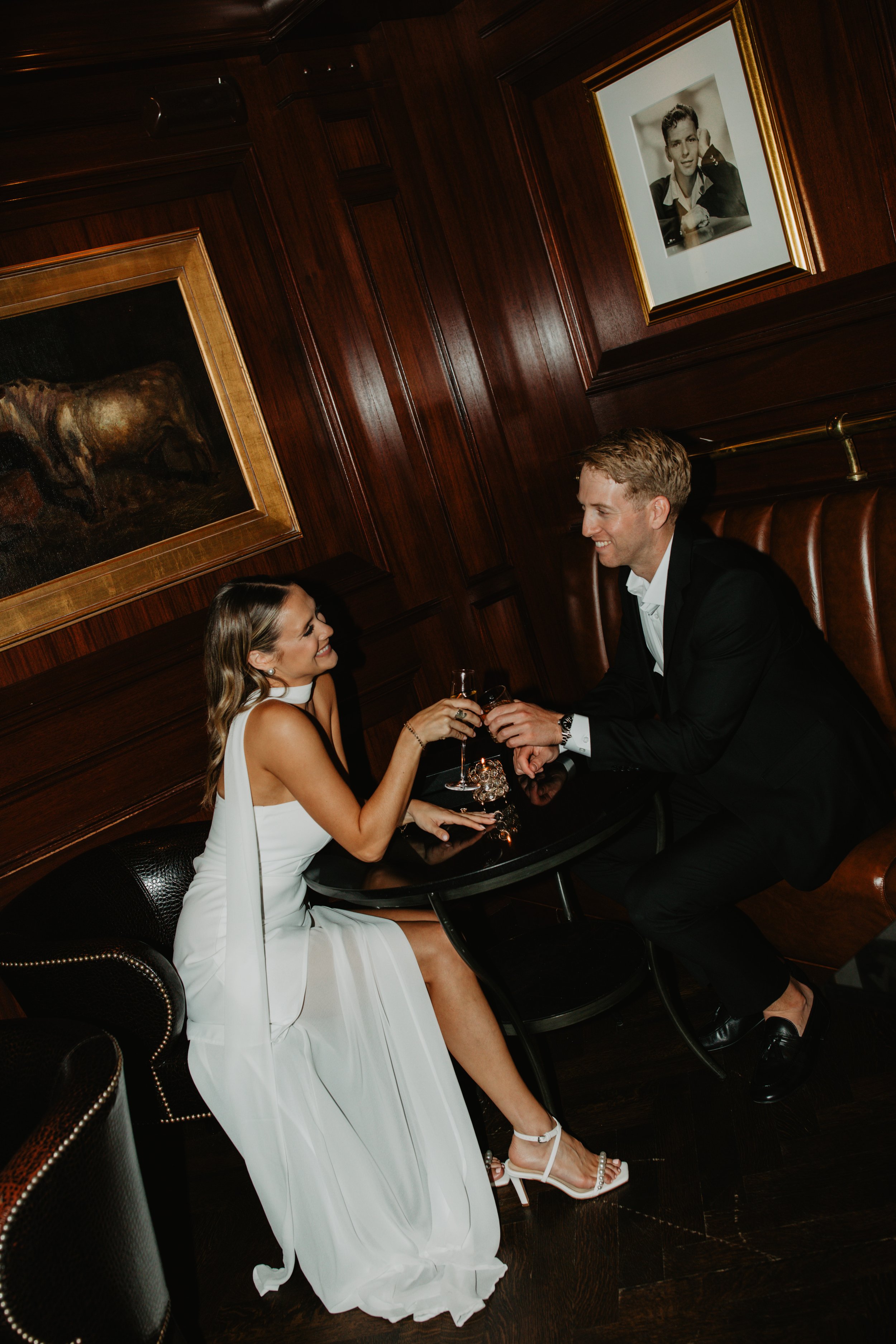 A woman and man dressed in formal attire sharing a toast at a restaurant table with dark wood paneling and framed artwork on the wall.