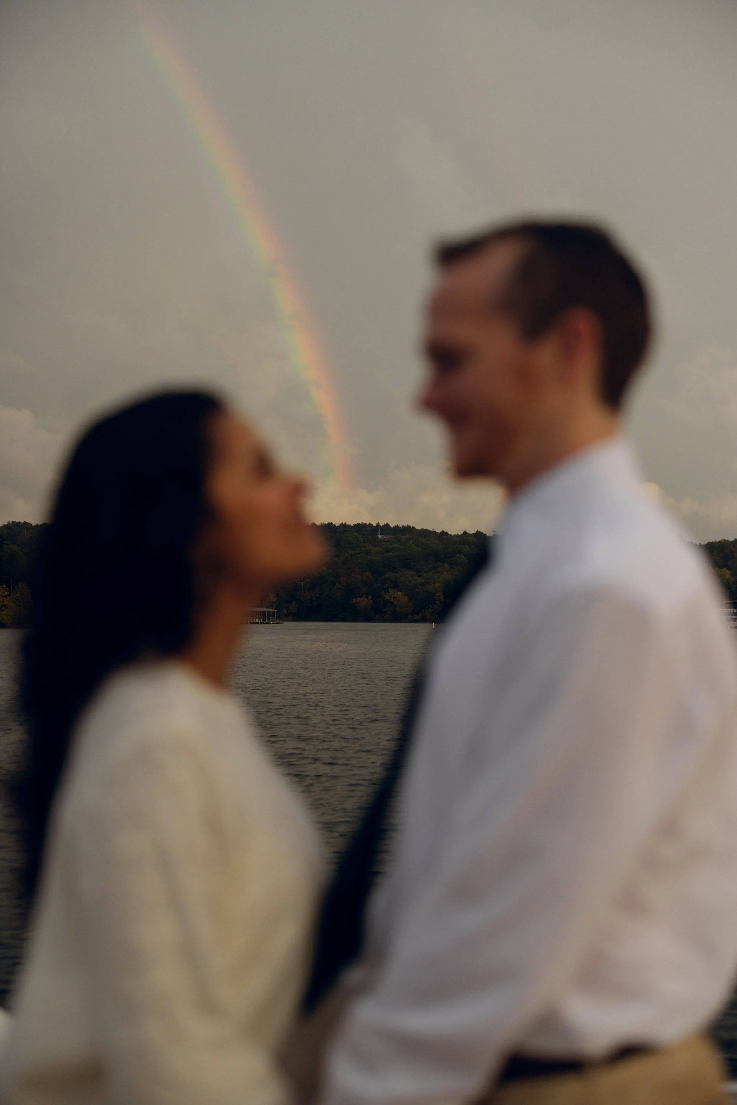 A blurry couple standing close together outdoors with a rainbow in the sky behind them.