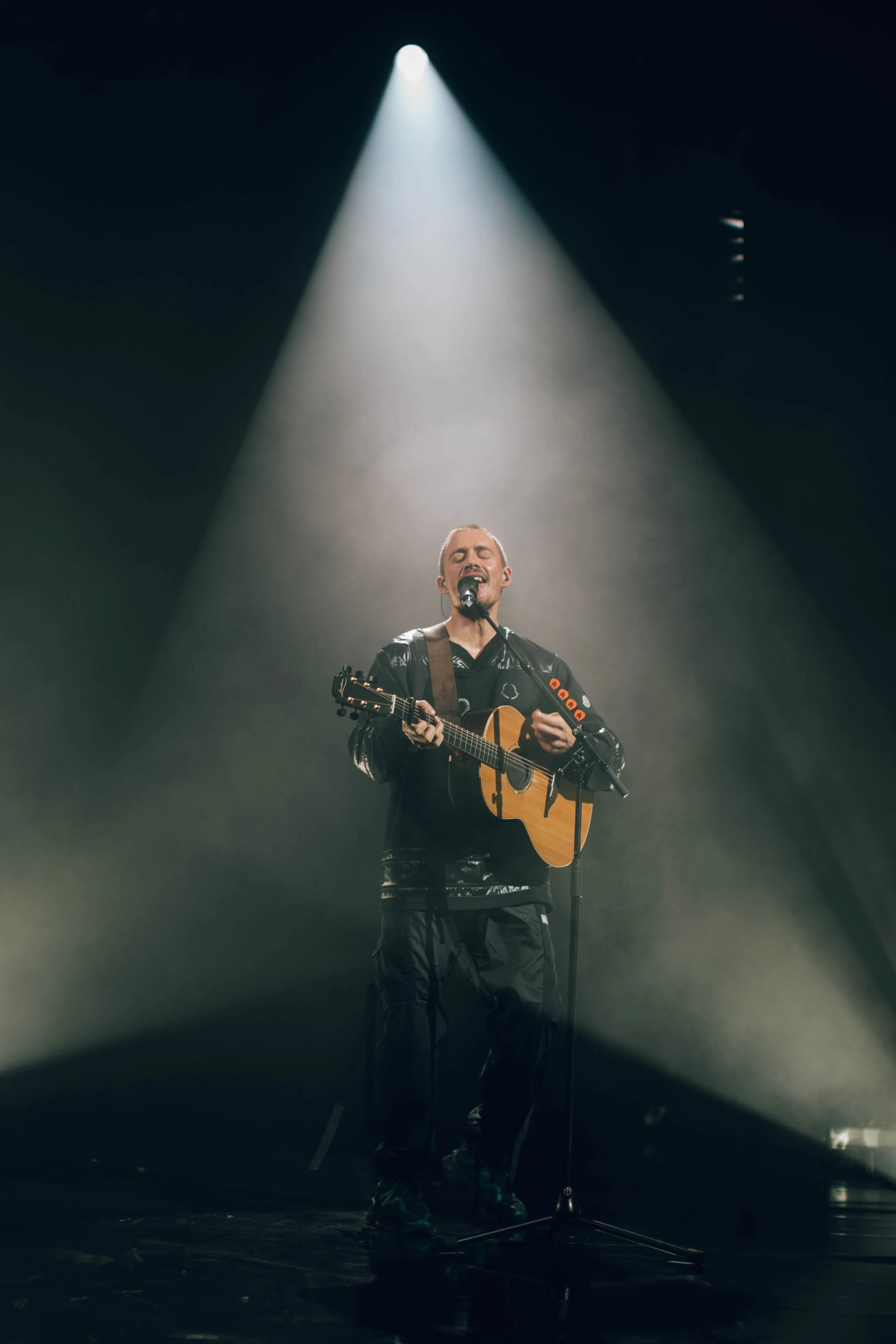 Dermot Kennedy singing and playing an acoustic guitar on stage under a spotlight.