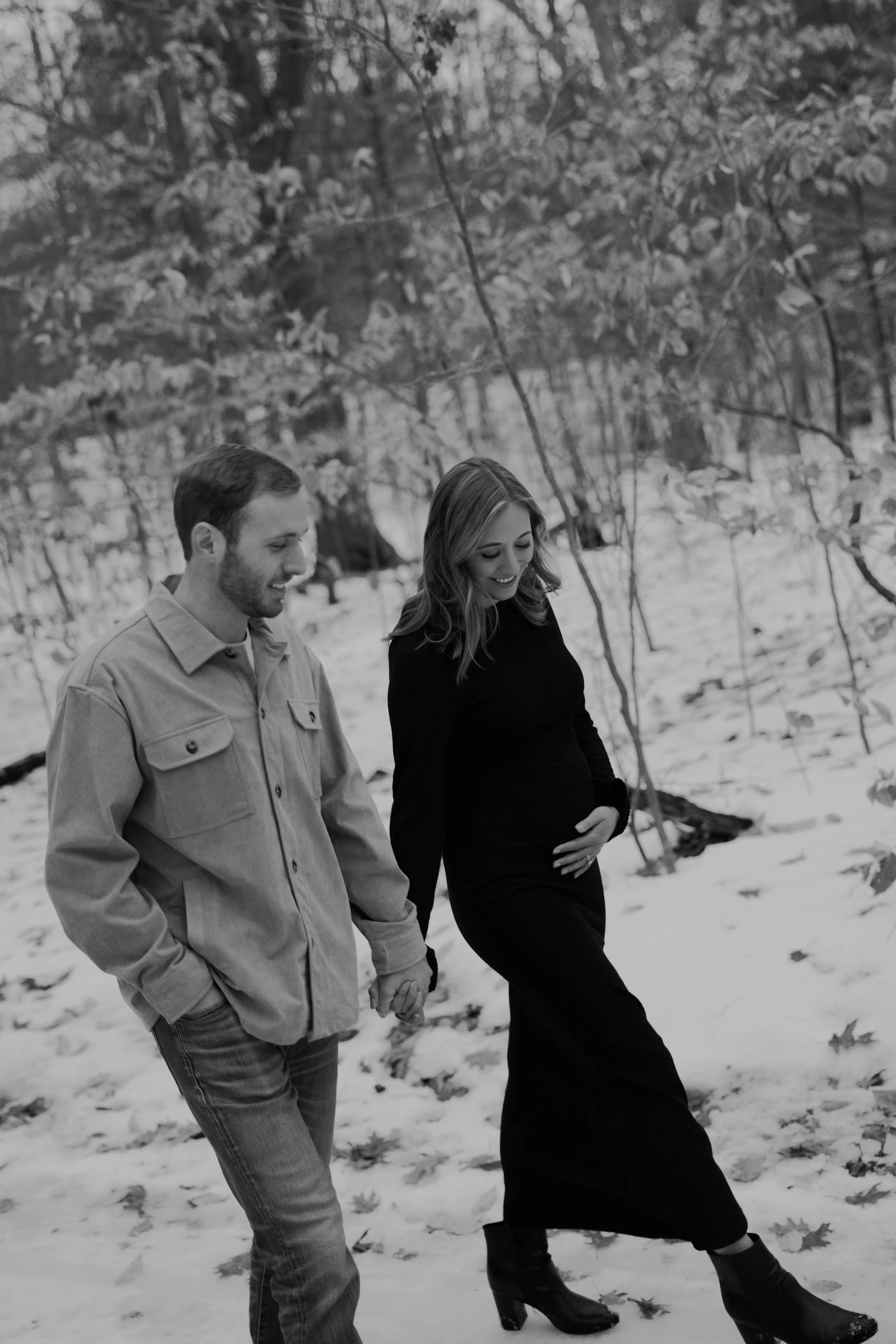 A black and white photo of a man and a pregnant woman walking hand in hand through a snowy forest, smiling and looking down.