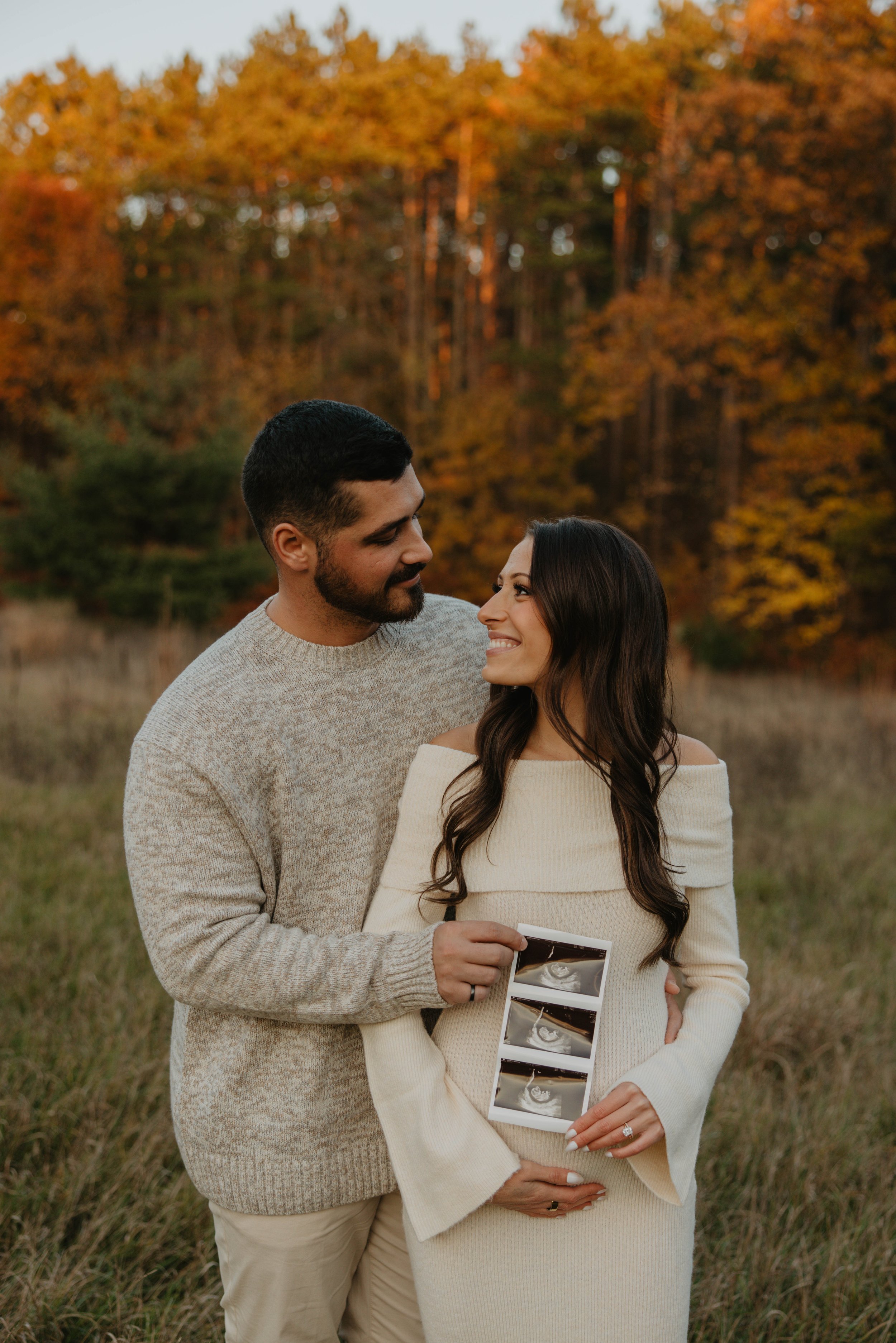 A couple standing in a field during fall, holding ultrasound pictures with trees in the background. The woman is pregnant and smiling at the man, who is looking at her lovingly.