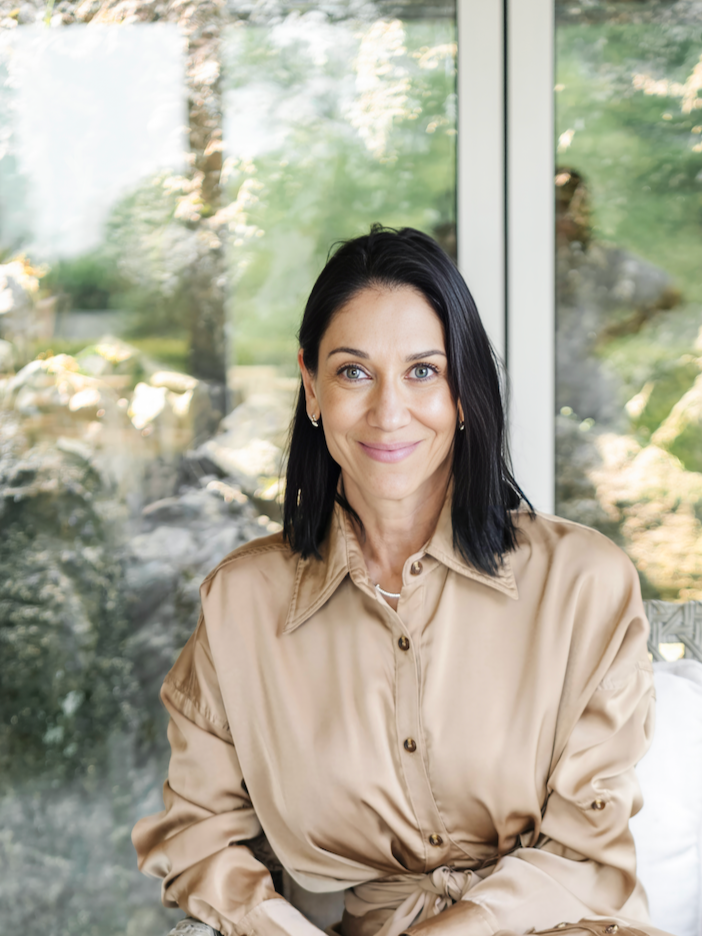 A woman with black hair, blue eyes, and earrings, wearing a beige blouse, sitting indoors near a glass window with greenery outside.