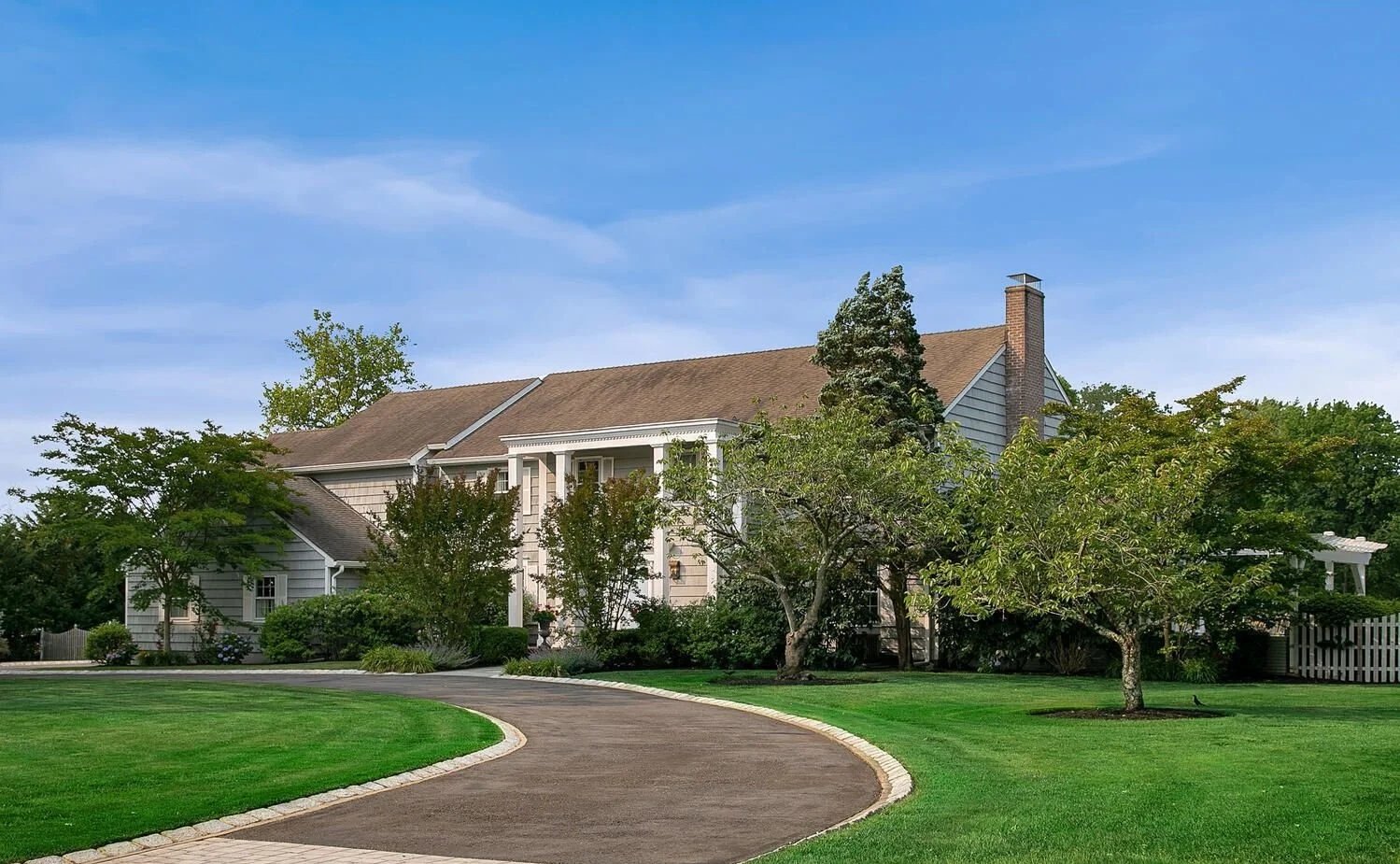 A large house with a driveway surrounded by well-maintained lawn and trees, under a clear blue sky.