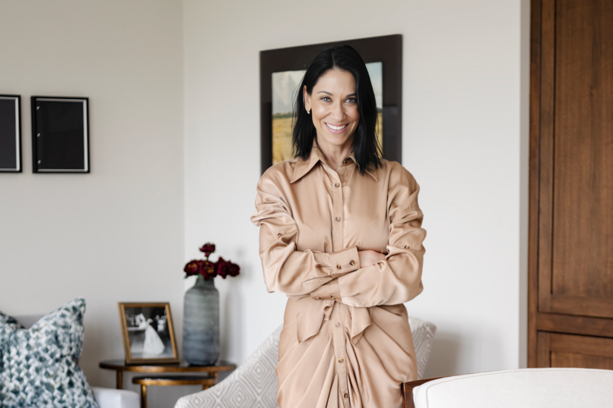 A woman with black hair and a beige satin shirt dress, smiling with arms crossed, standing in a living room with a framed picture, flowers in a vase, and patterned pillows.