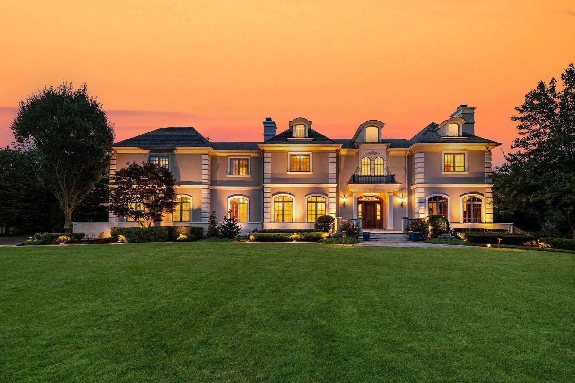 A large, elegant house illuminated at dusk. The house has multiple stories with a dark roof, large windows, and a grand entrance with steps and lighting. There are trees and a well-kept lawn in front of the house.