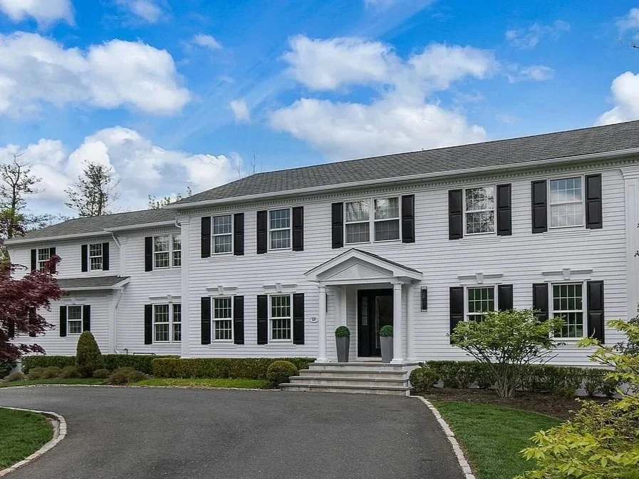 A large white two-story house with black shutters, a covered front porch, and a neatly landscaped yard under a blue sky with clouds.