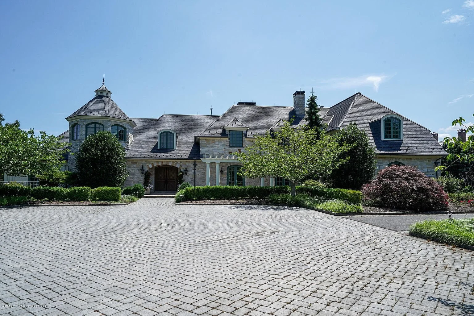 Large stone house with a turret, multiple chimney stacks, and a paved driveway surrounded by landscaped gardens with trees and bushes.
