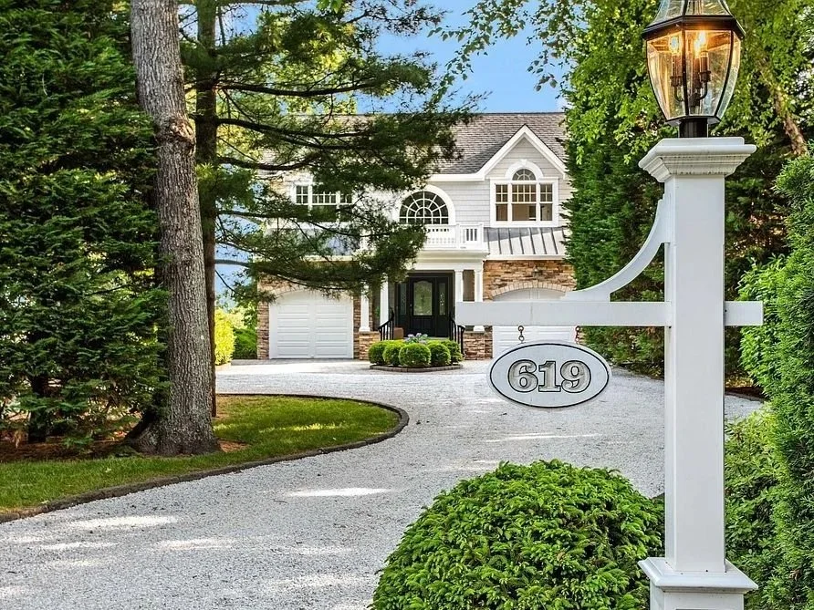 A white mailbox with an oval house number sign reading "619" in front of a two-story house with white siding, large arched window, and attached garage, surrounded by greenery and trees, with a gravel driveway and a lamp post.