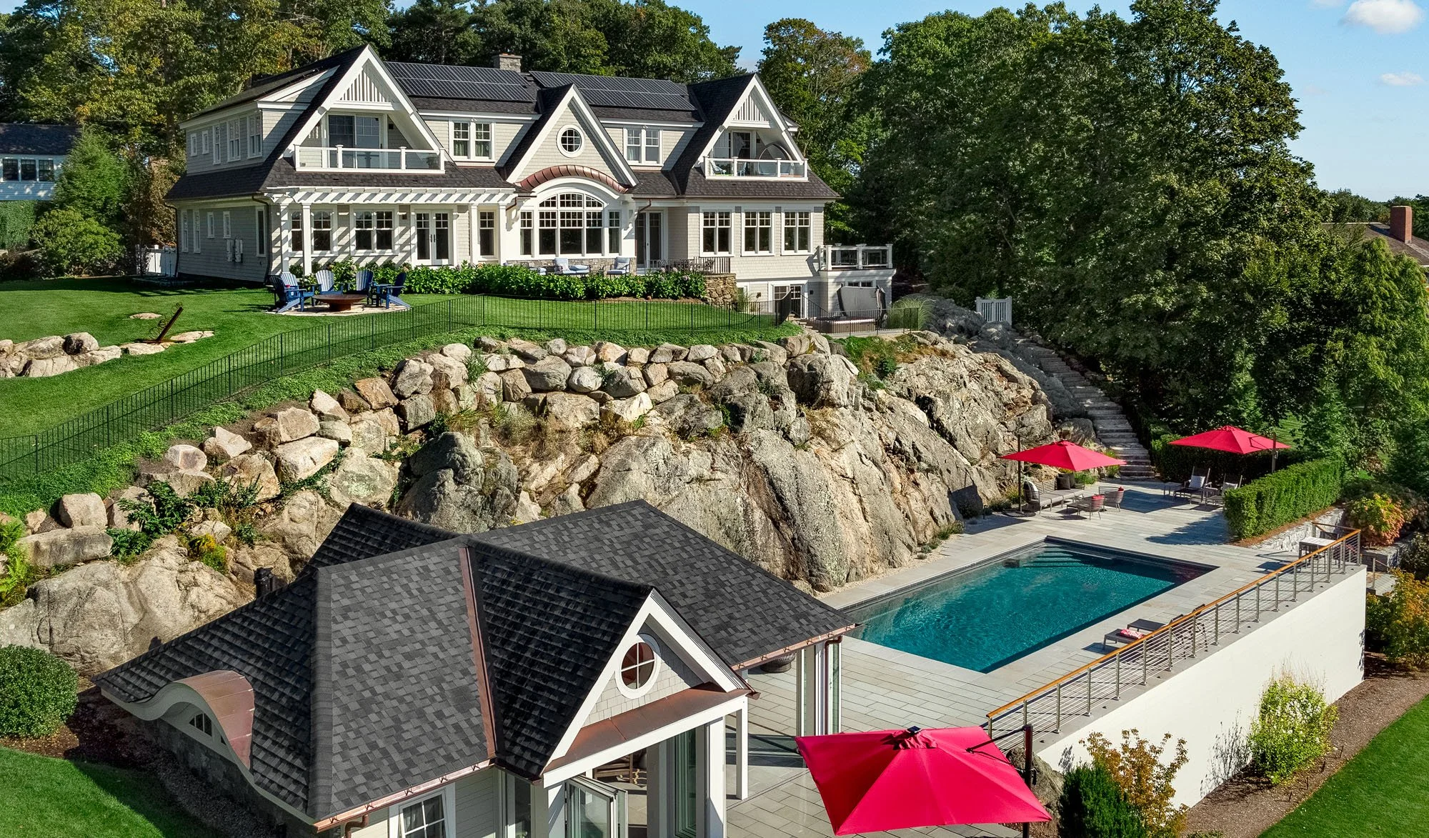 A large luxury house on a rocky hill with a backyard featuring a swimming pool, outdoor seating areas with red umbrellas, and a well-maintained grassy lawn with trees.