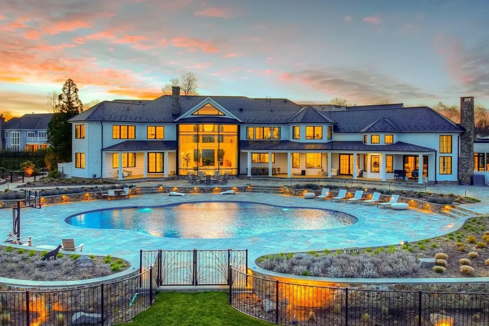 A large, modern house with many windows illuminated from inside, overlooking a backyard swimming pool. The pool area is surrounded by chairs and a fence, with sunset sky in the background.
