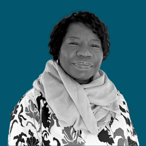 Portrait of a woman with short curly hair, wearing a patterned blouse and a light scarf, against a solid teal background.