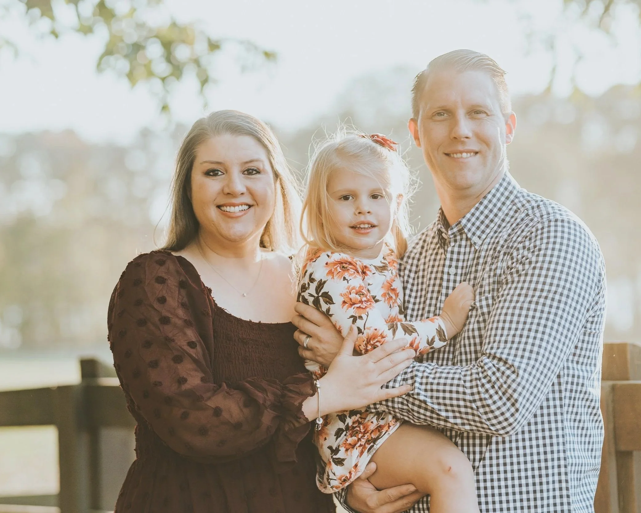 A family of three, a woman, a man, and a young girl, posing outdoors on a sunny day. The woman wears a dark dress, the man a checkered shirt, and the girl a floral dress. The man holds the girl, and the woman touches the girl’s arm.