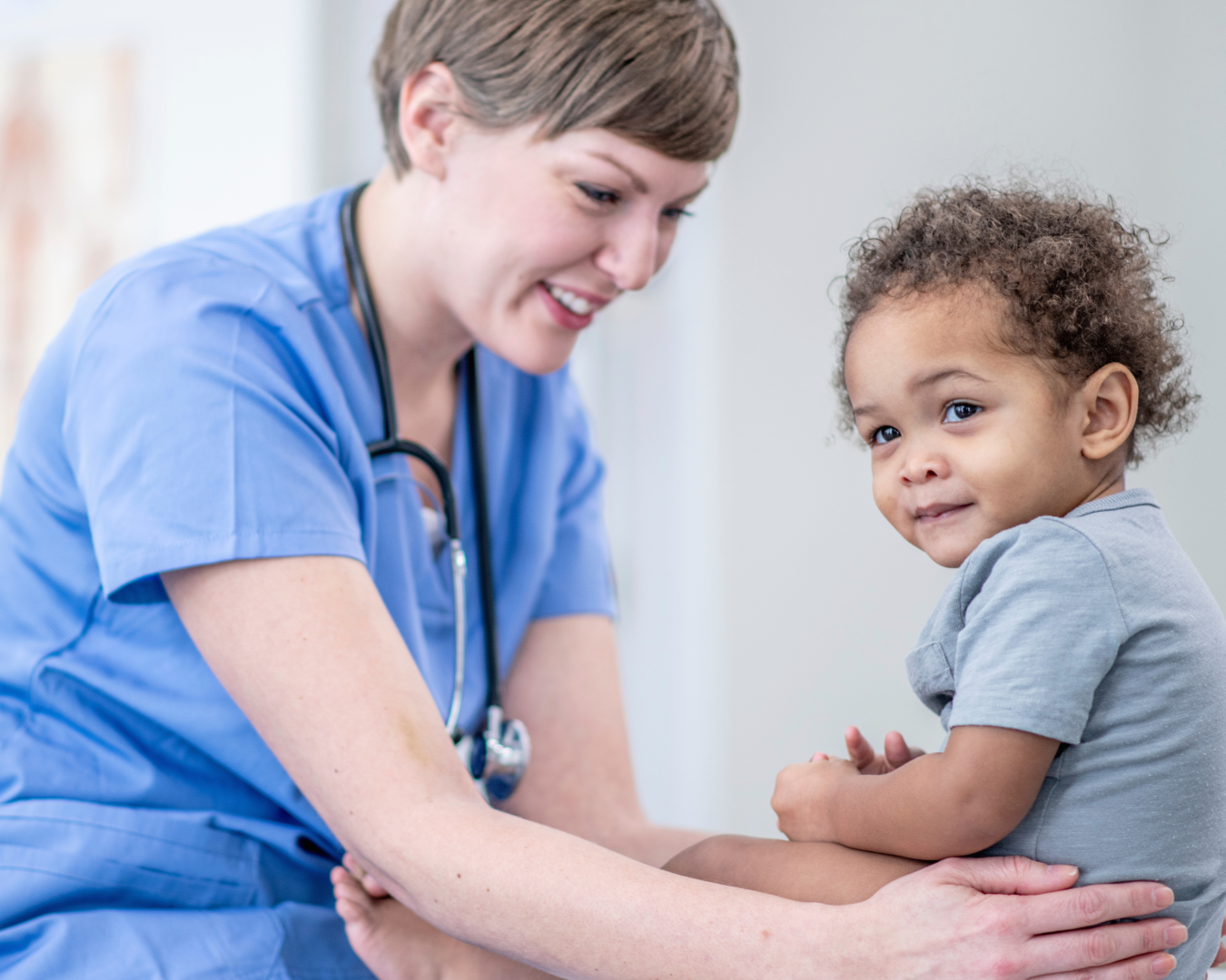 A healthcare worker in blue scrubs smiling at a young child who is sitting on her lap, looking at the camera.